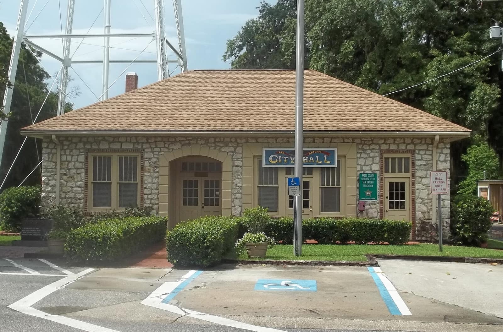 Small stone City Hall building with a hipped roof, arched entrance and landscaped shrubs on a sunny day.