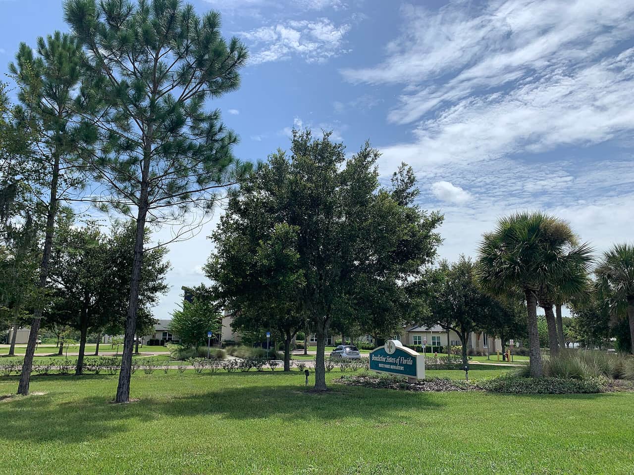 Green lawn, tall pine and oak trees, and a sign reading Holy Name Monastery under blue sky with clouds.