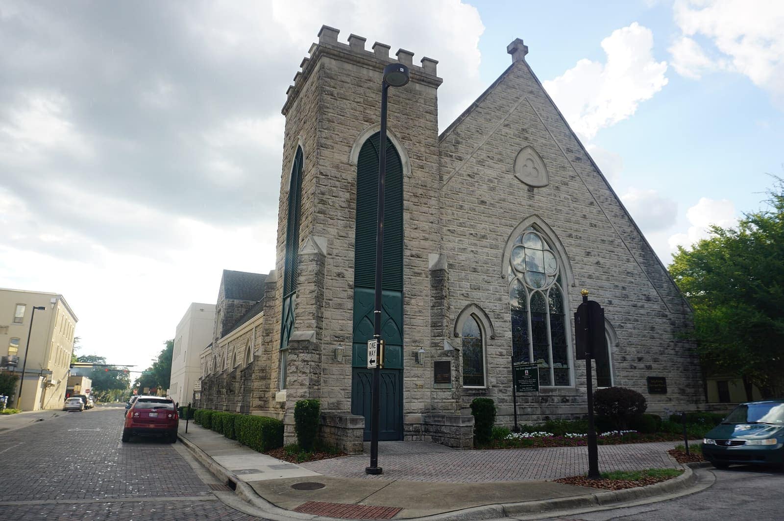 A historic stone Gothic-style church with a square tower and pointed windows stands beside a city street under cloudy skies.