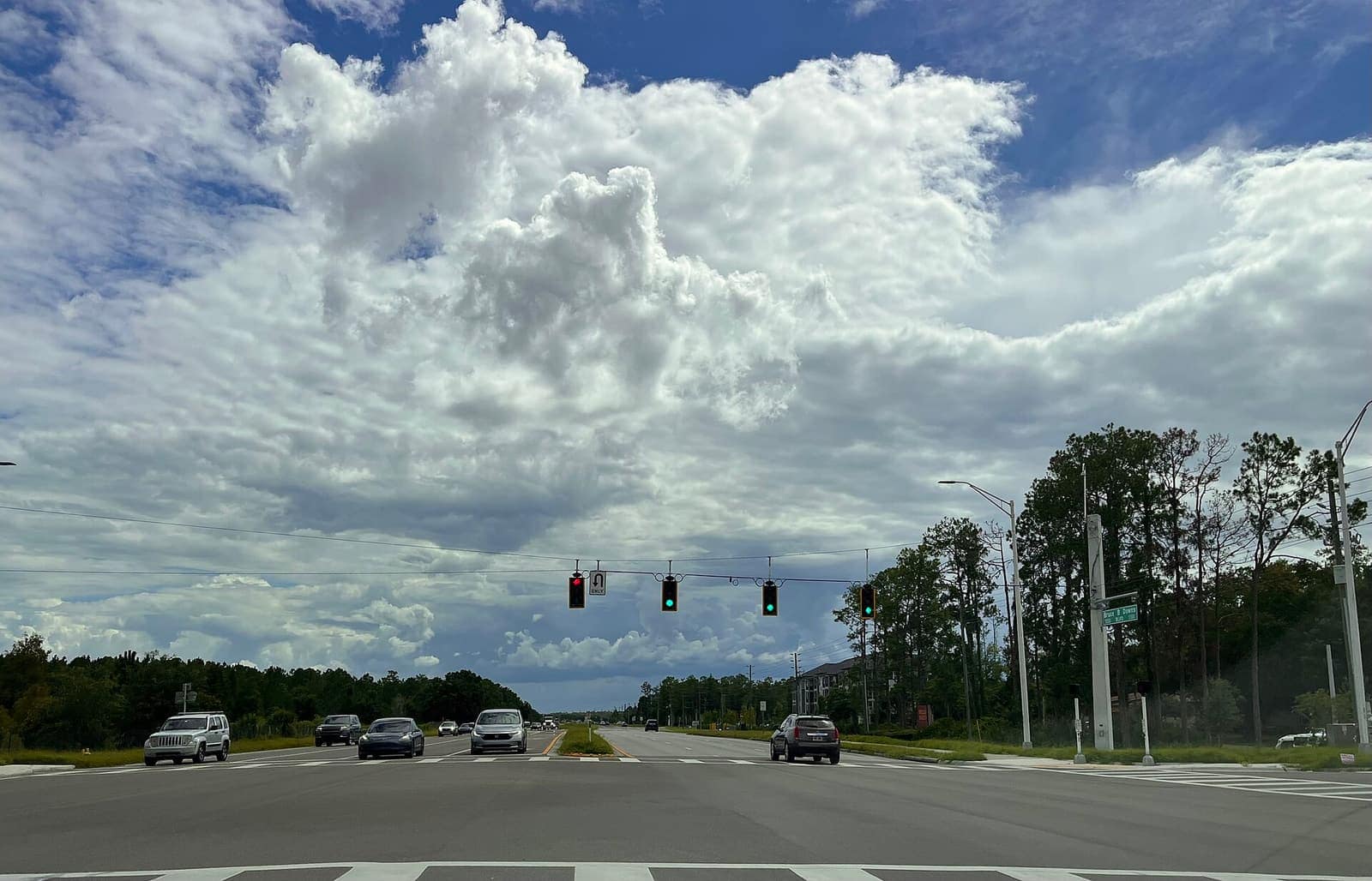 A signalized intersection under large cumulus clouds with cars waiting and traffic lights hanging overhead.