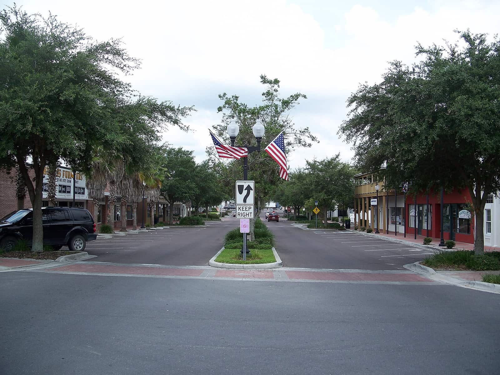 Tree-lined downtown street with two American flags, a keep-right sign and storefronts under a cloudy sky.