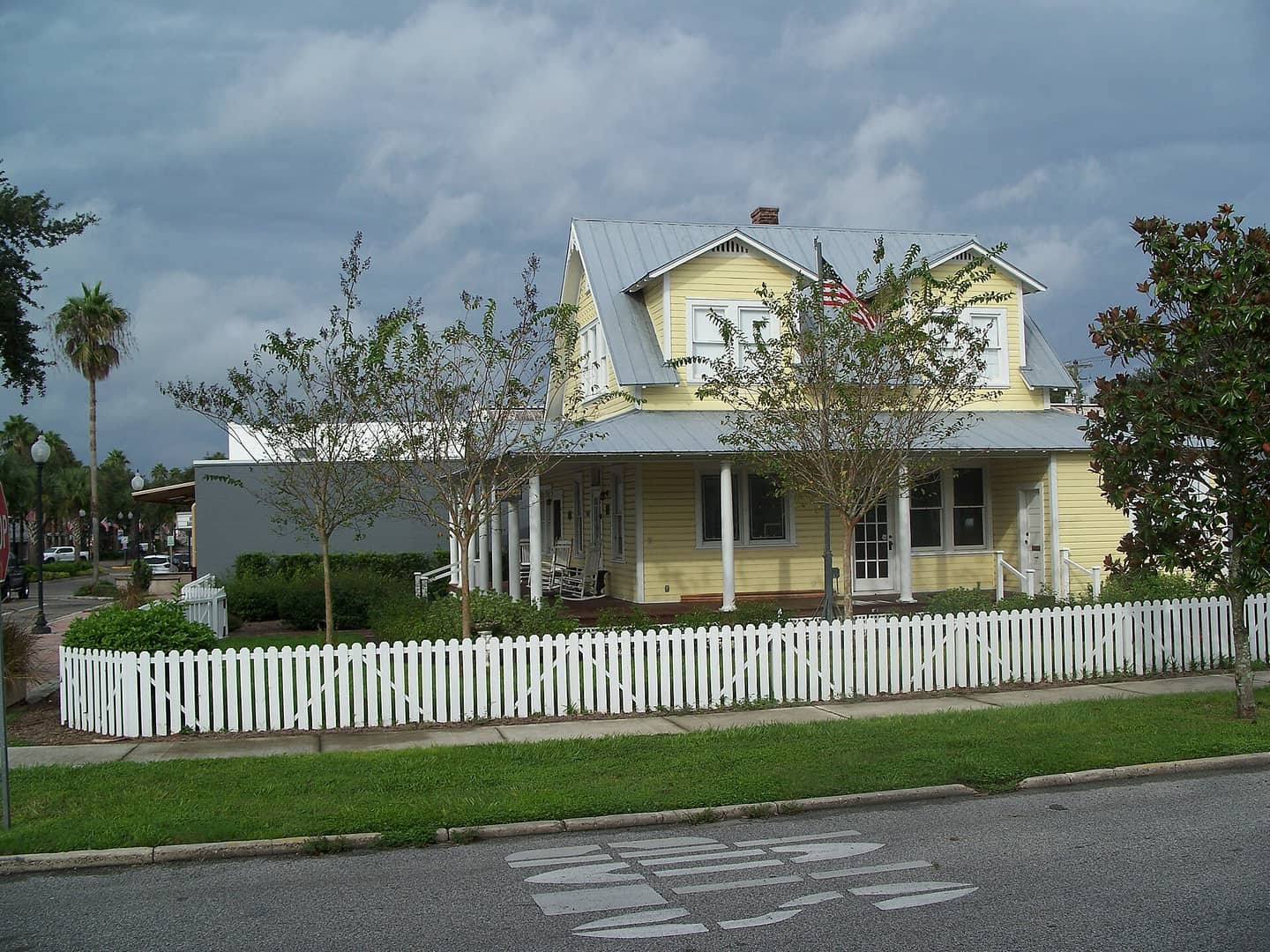 Yellow two-storey wood house with white picket fence, front porch and metal roof under cloudy sky.