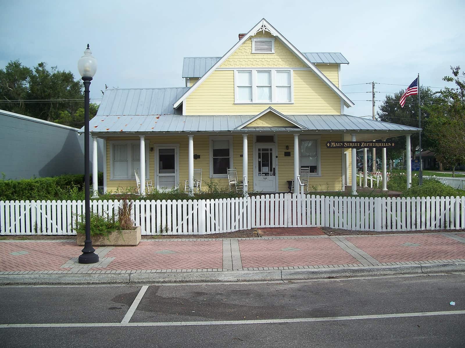 Yellow historic house with white picket fence, covered porch, lamp post and American flag on a sunny street.