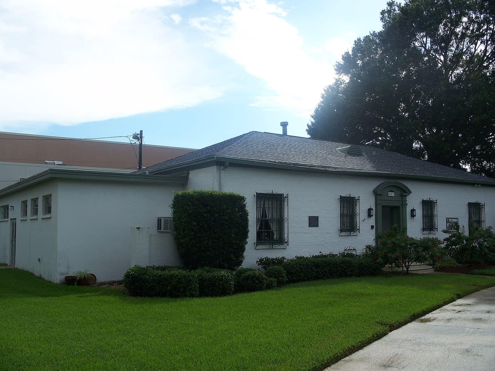 White stucco single-story building with barred windows, green lawn, and clipped hedges beside a driveway.