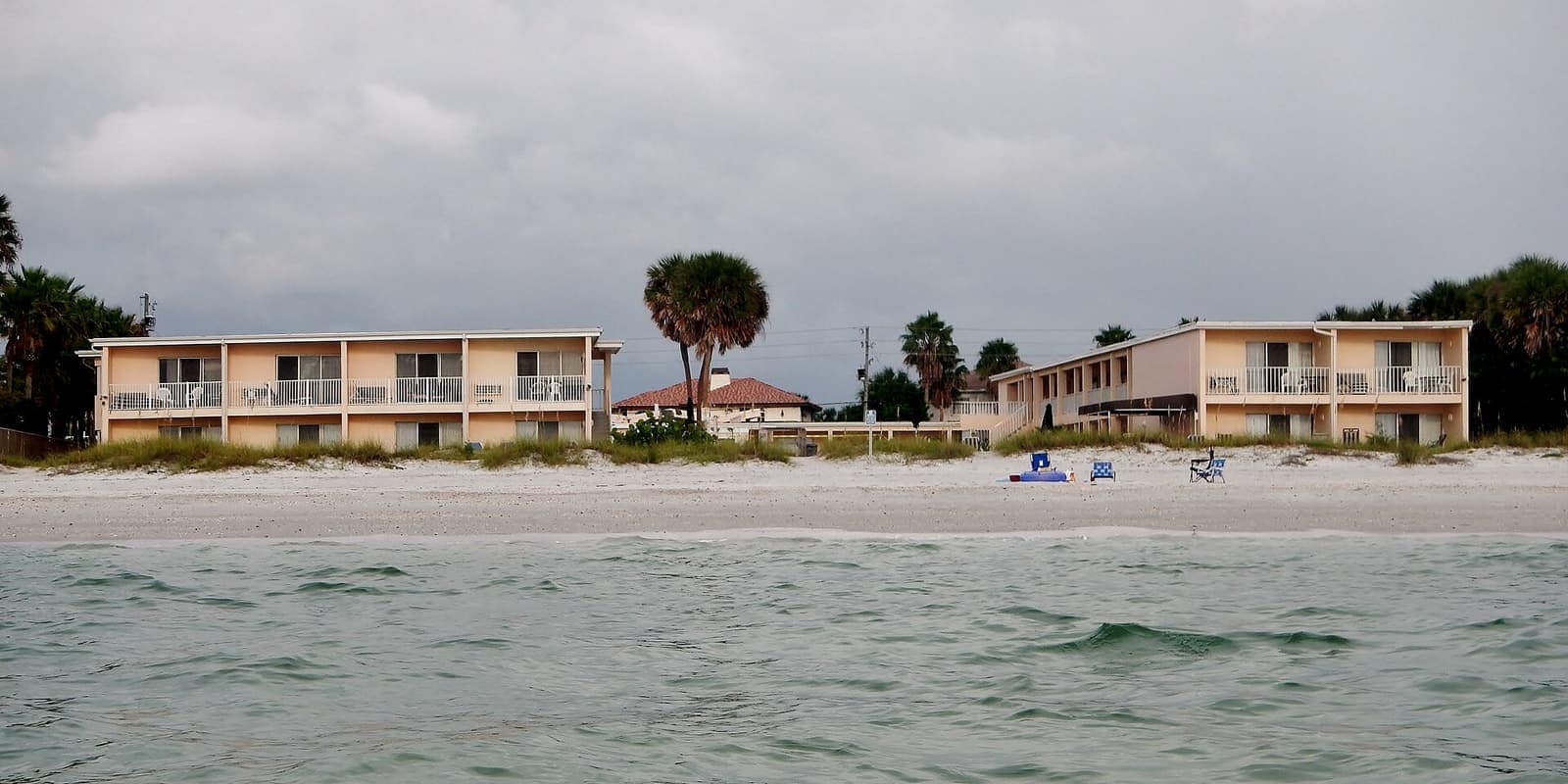 Two-story beachfront motel buildings above a white sand beach under overcast sky.