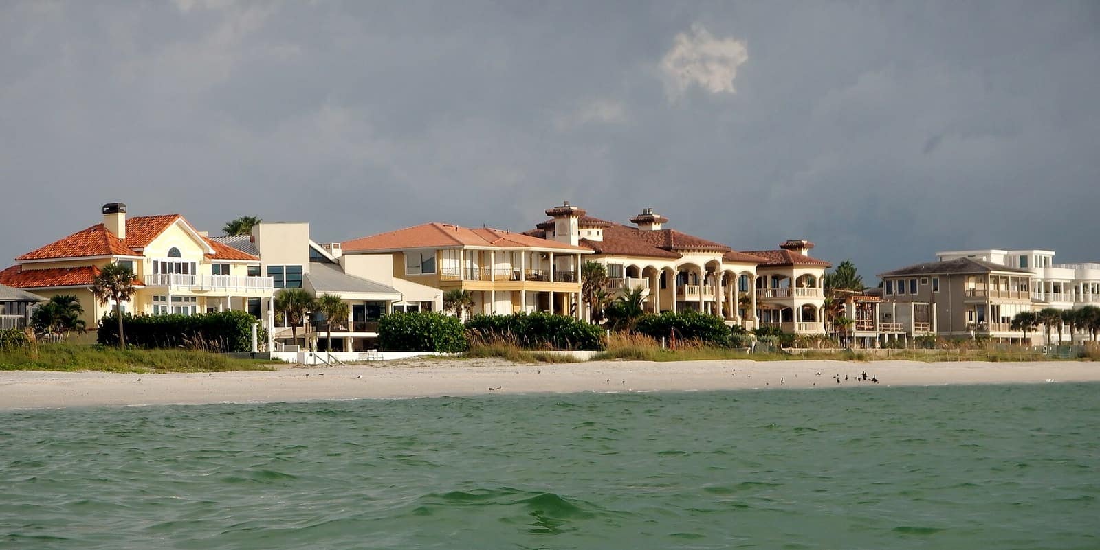Large tile-roofed beachfront homes line a sandy shore beside green water under cloudy sky.