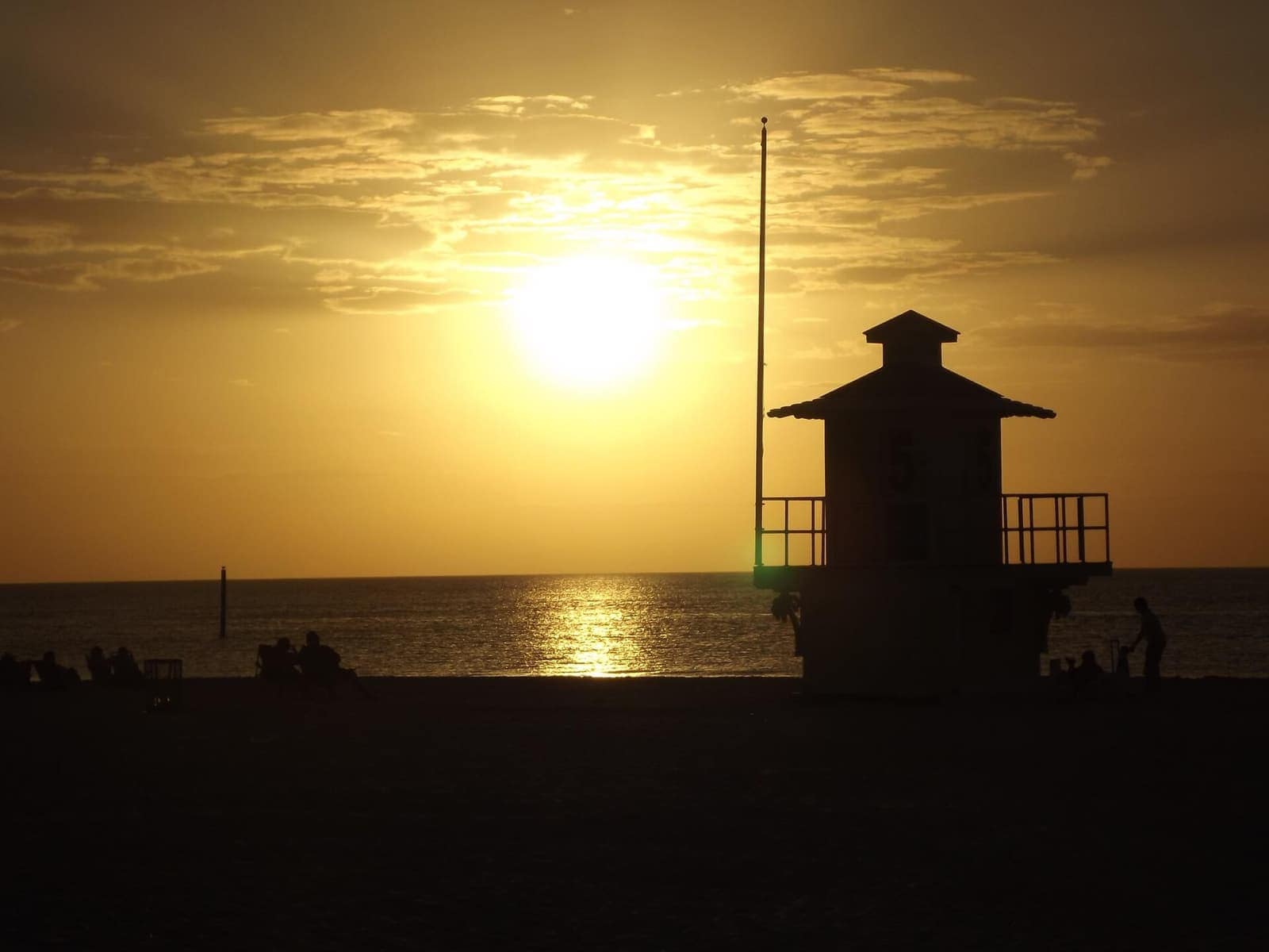Silhouetted lifeguard tower and small figures on a beach at sunset over calm water.