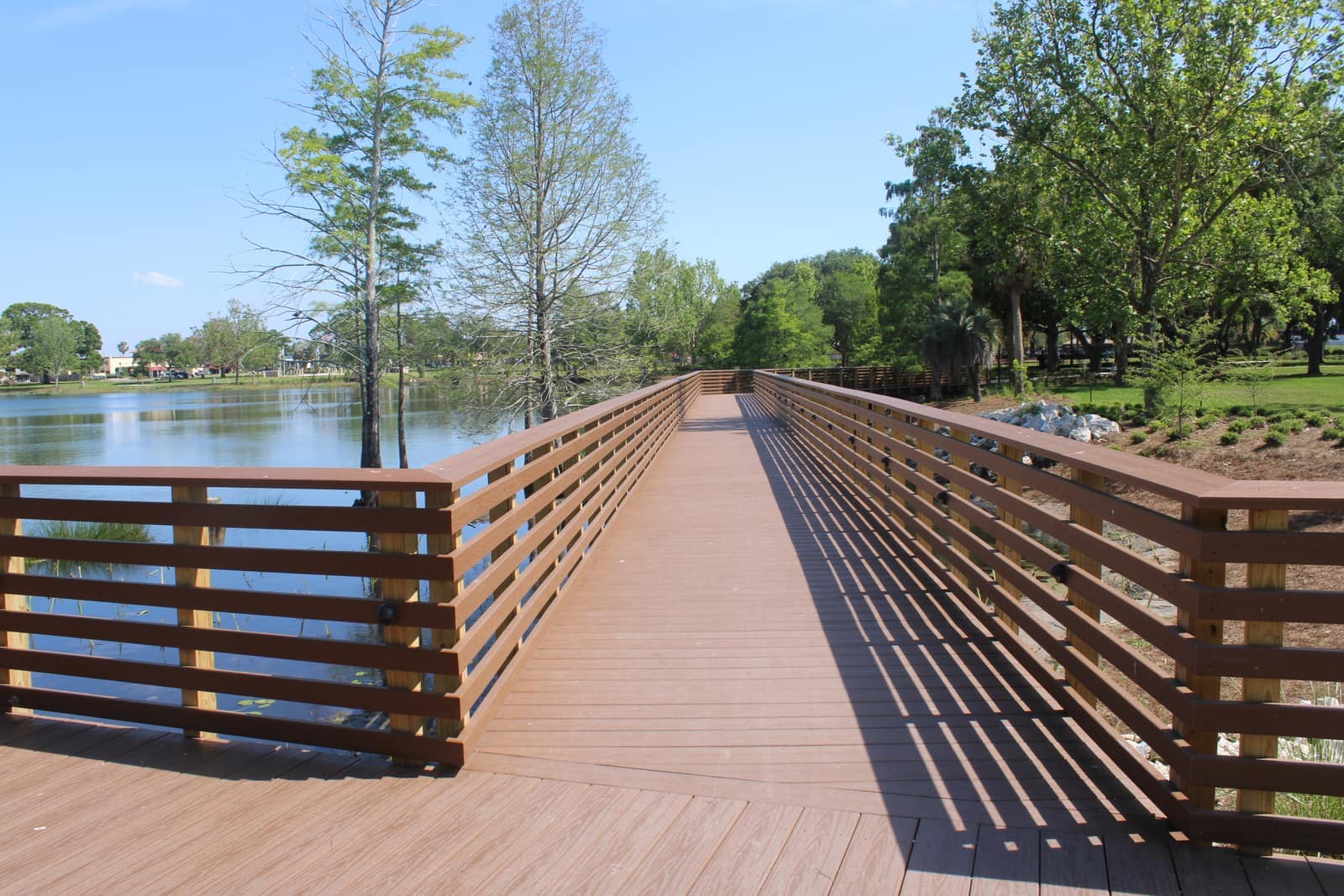 Wooden boardwalk with railings extending across a calm lake toward trees under a clear blue sky.