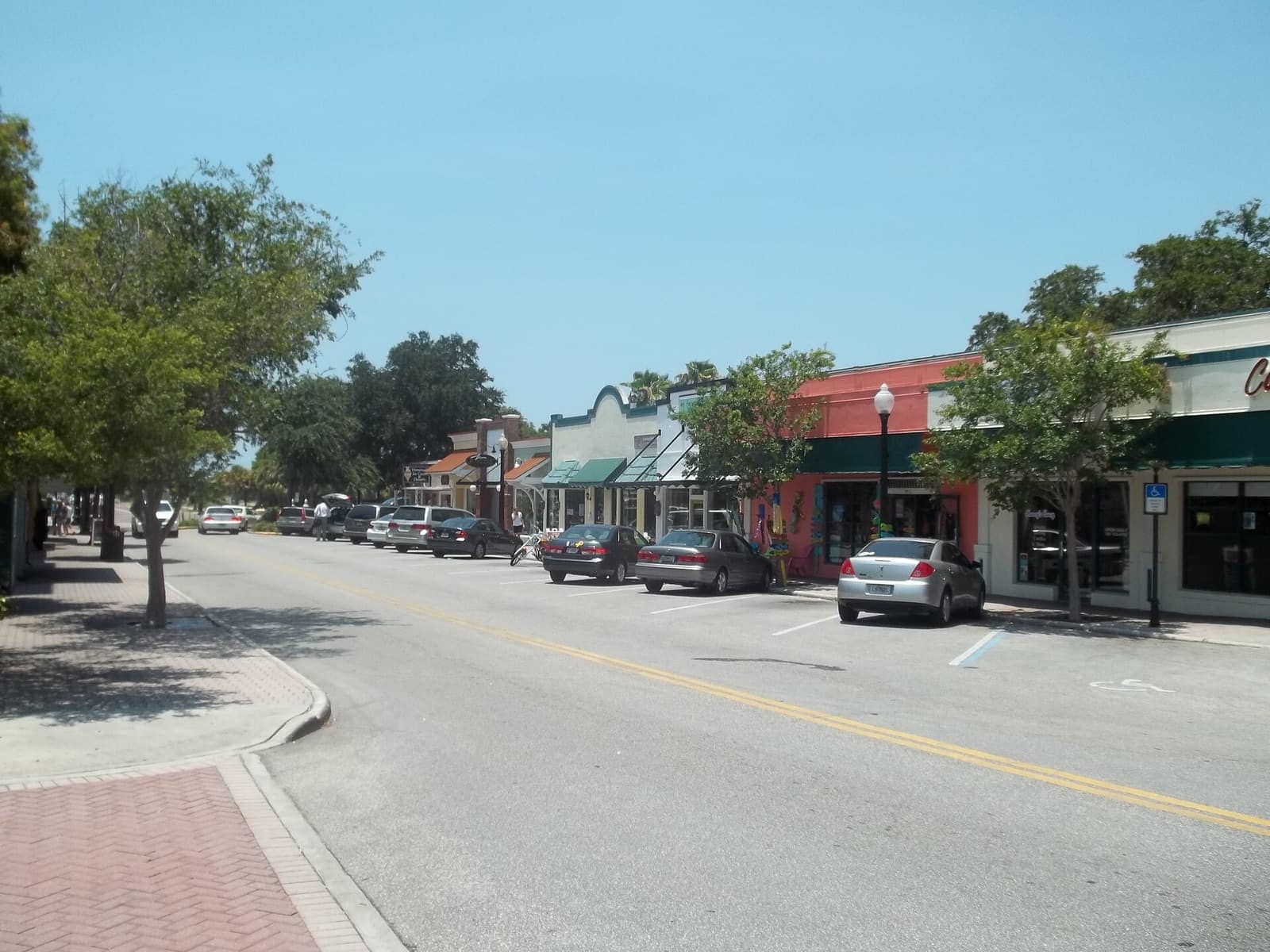 Tree-lined downtown street with parked cars and colorful shopfronts under a clear blue sky.