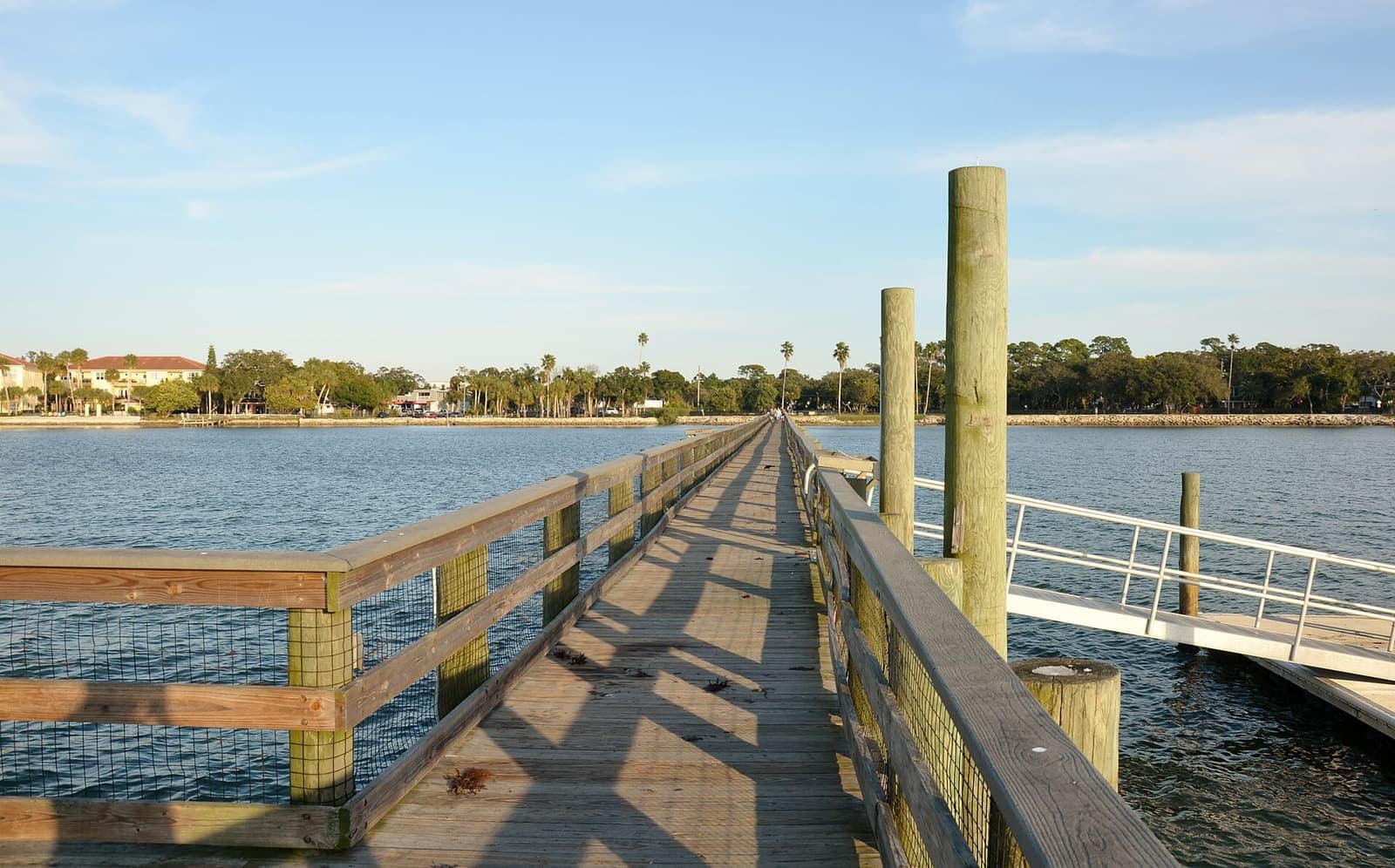 Wooden fishing pier with railings stretching over calm water toward palm trees and shoreline buildings.