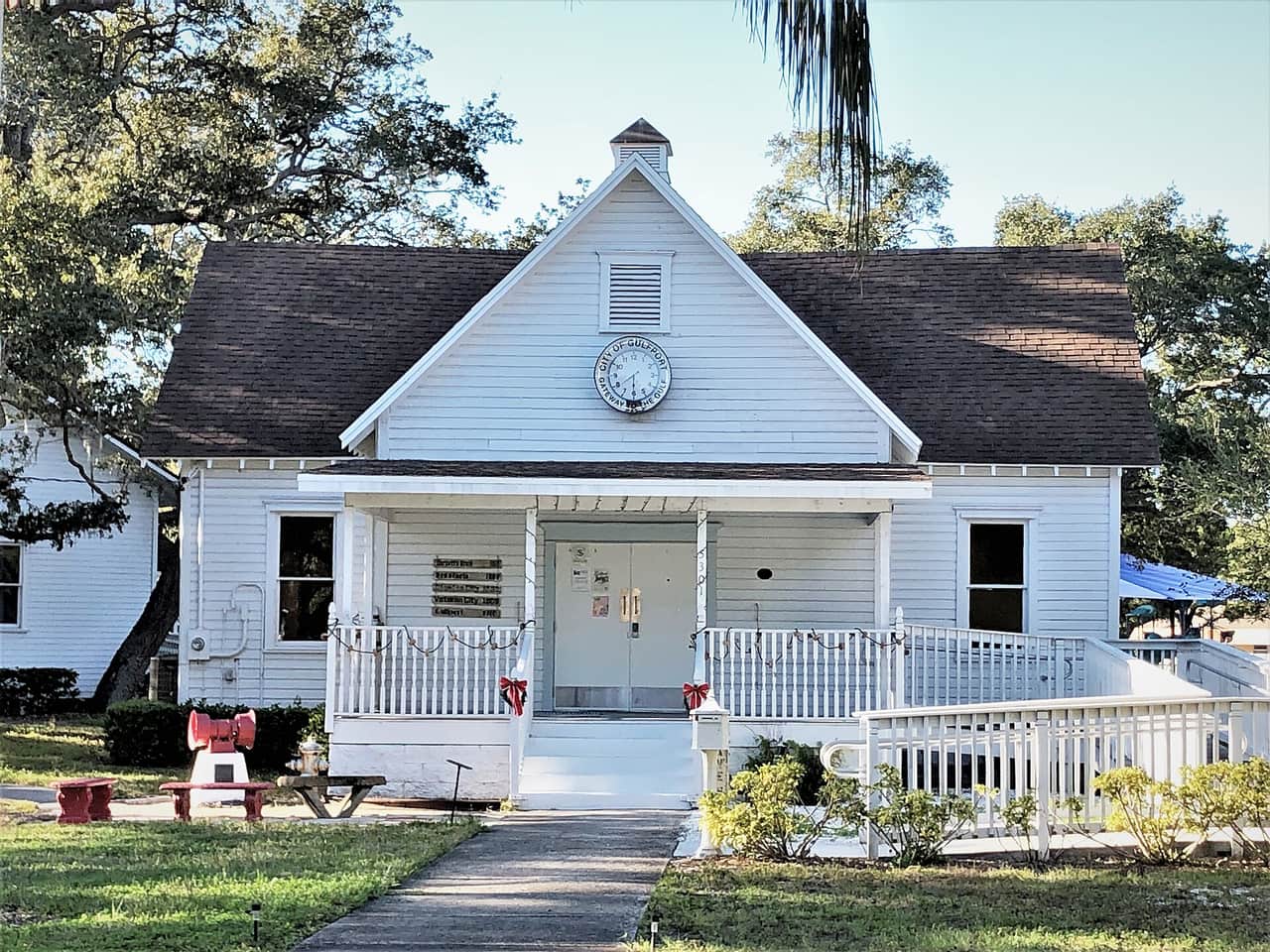 A small white wooden building with a gabled roof, clock, and porch under tall trees in daylight.