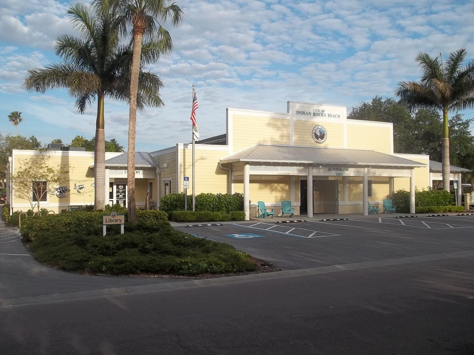 Yellow municipal building with a flagpole, palm trees, and a parking lot under a partly cloudy sky.