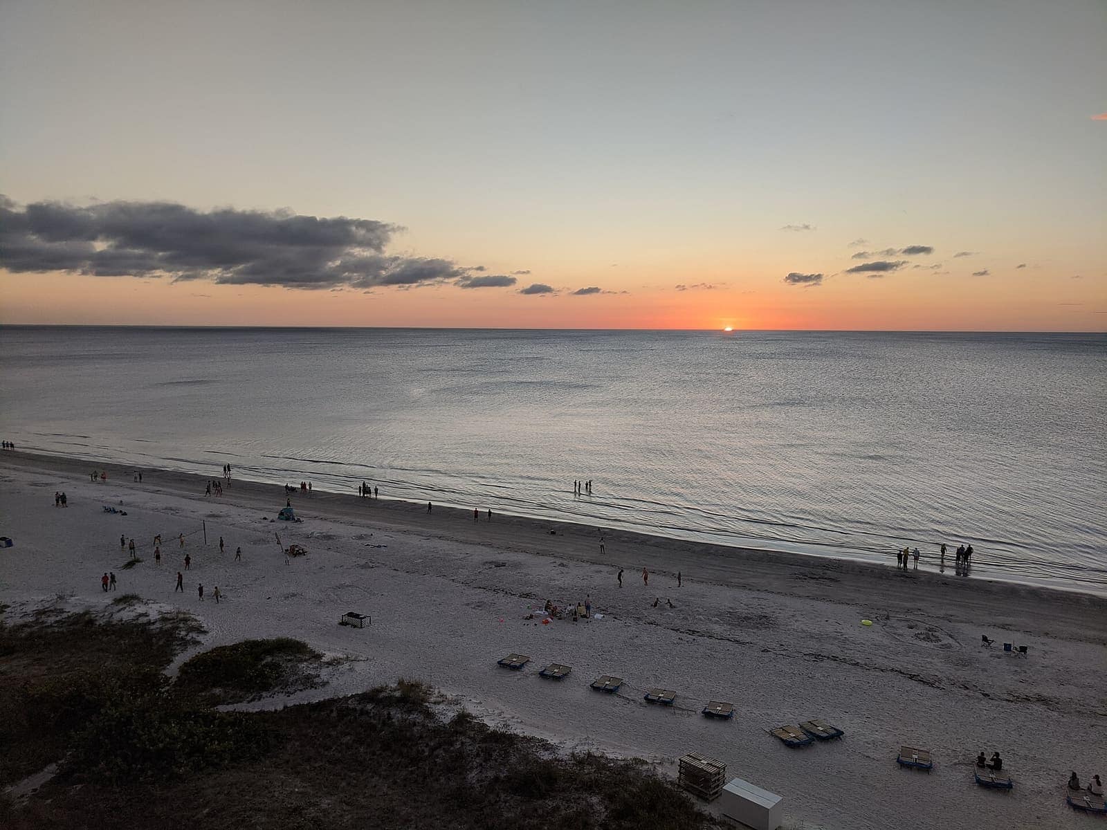Sun sets over a calm Gulf of Mexico with small distant figures on a sandy beach.