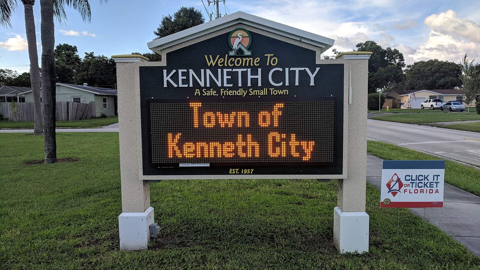 Dark welcome sign reading Welcome To Kenneth City above an orange Town of Kenneth City message board.