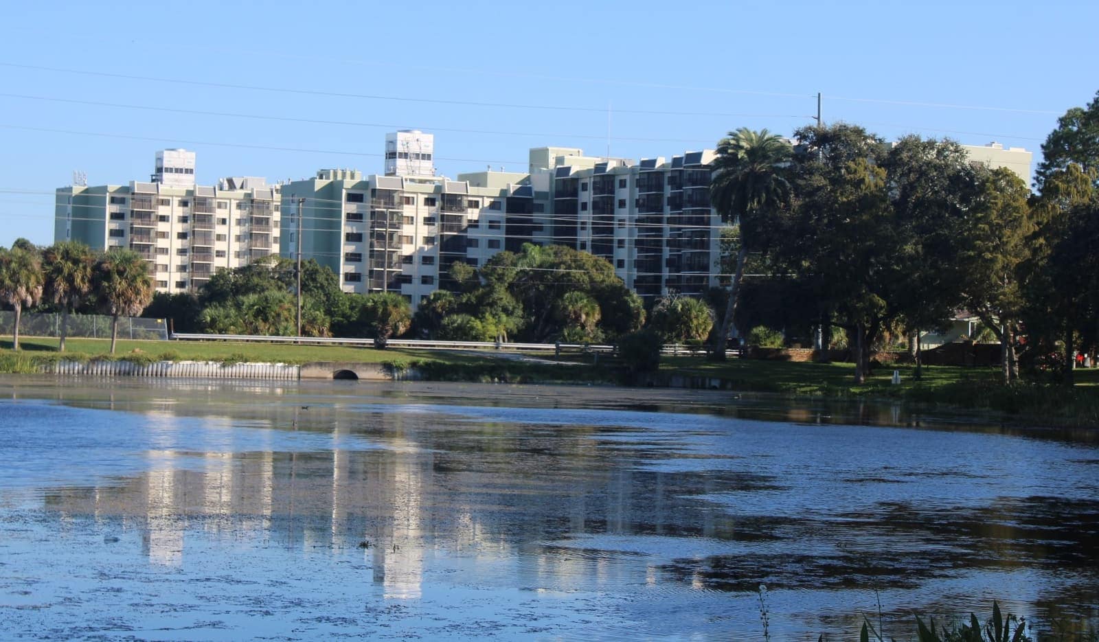 A calm lake reflects a row of pale multi-story apartment buildings under a clear blue sky.