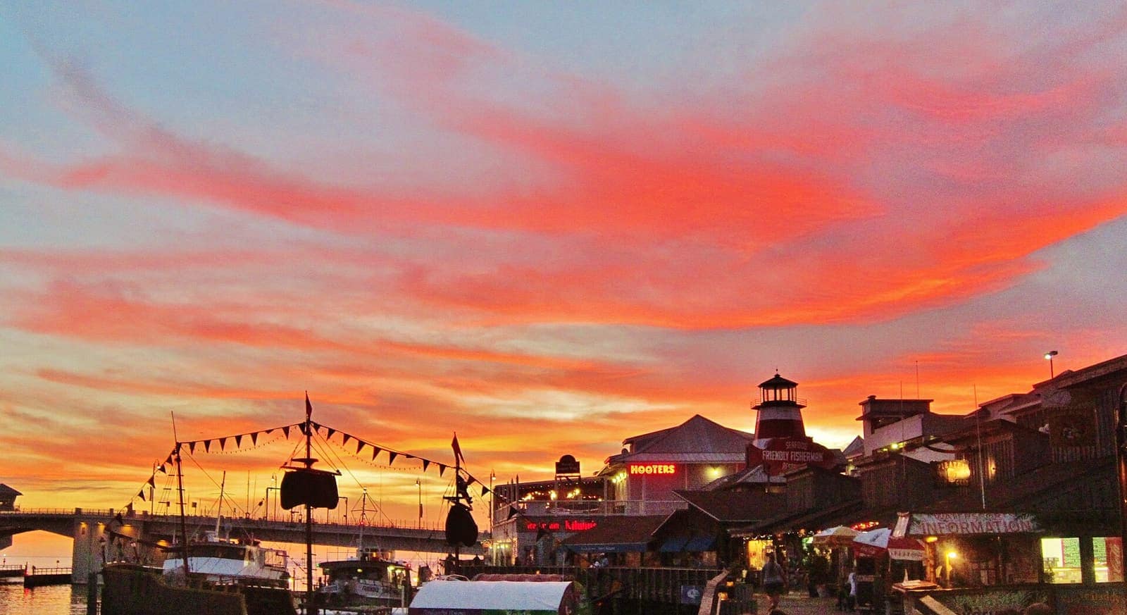 Bright red and pink clouds over a waterfront village with a tower, bridge, and moored boats.