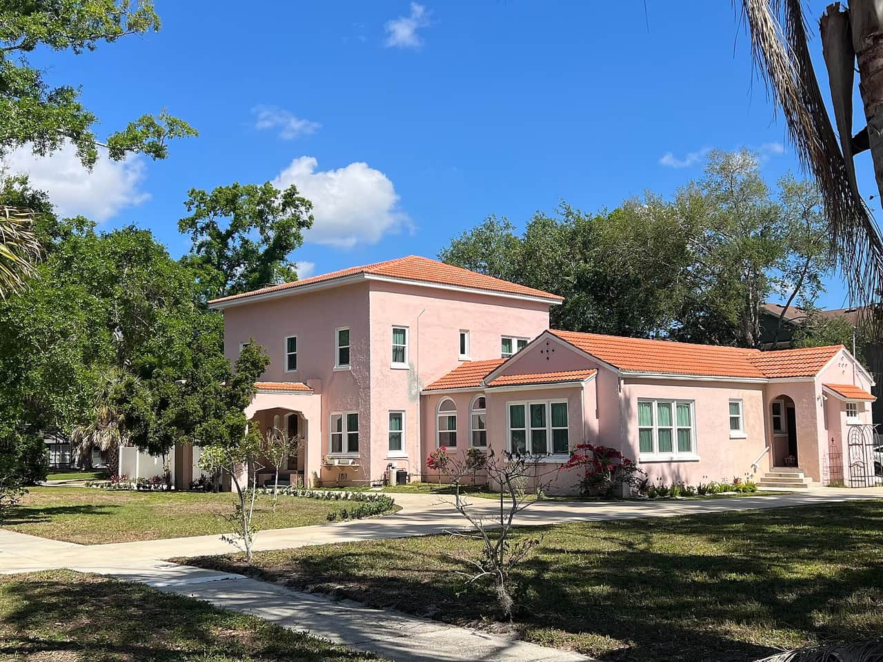 Pink stucco Mediterranean-style house with terracotta roof and green lawn under blue sky.