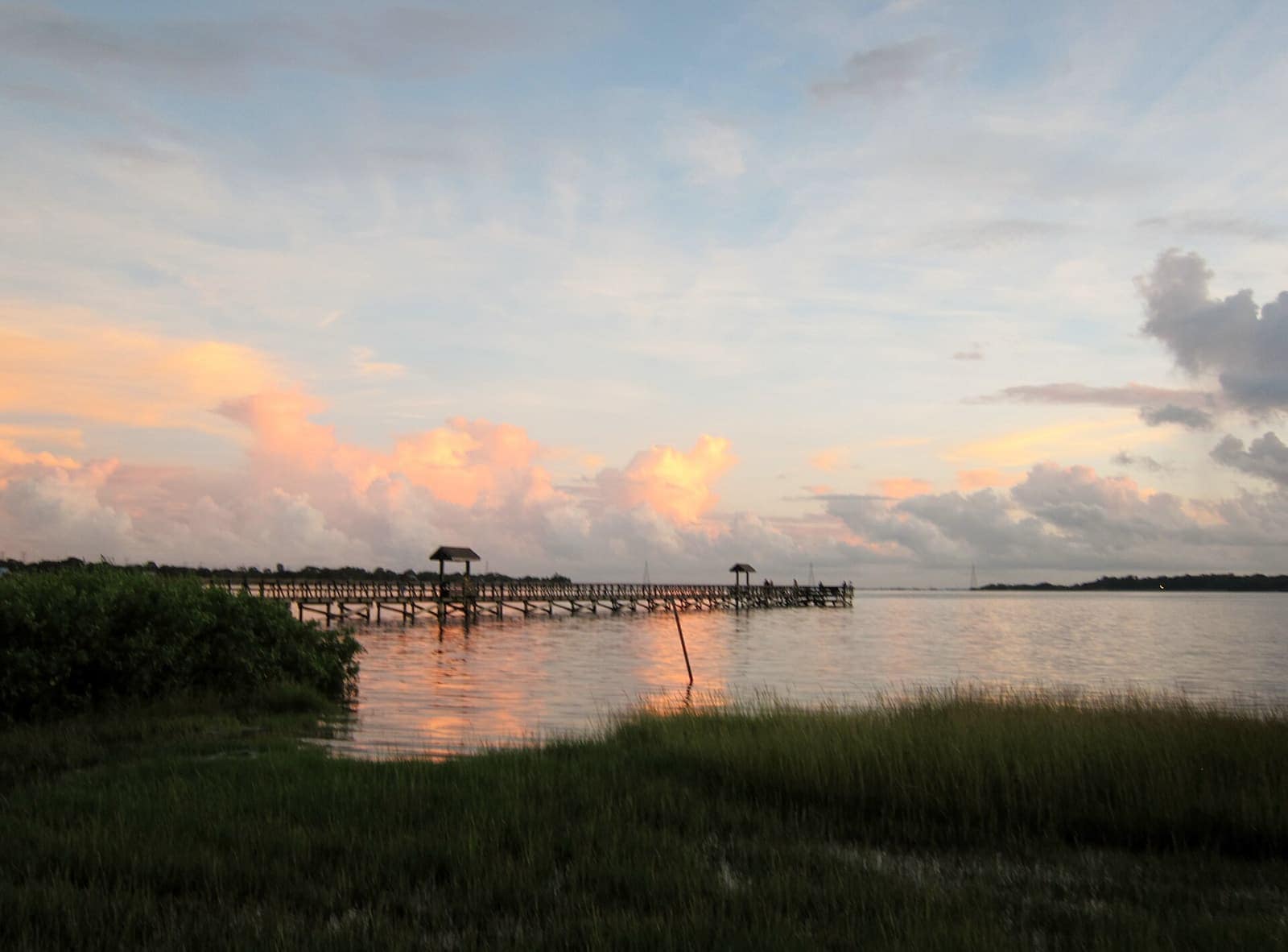 Wooden fishing pier extending over calm water at sunset with pink clouds and marsh grass.