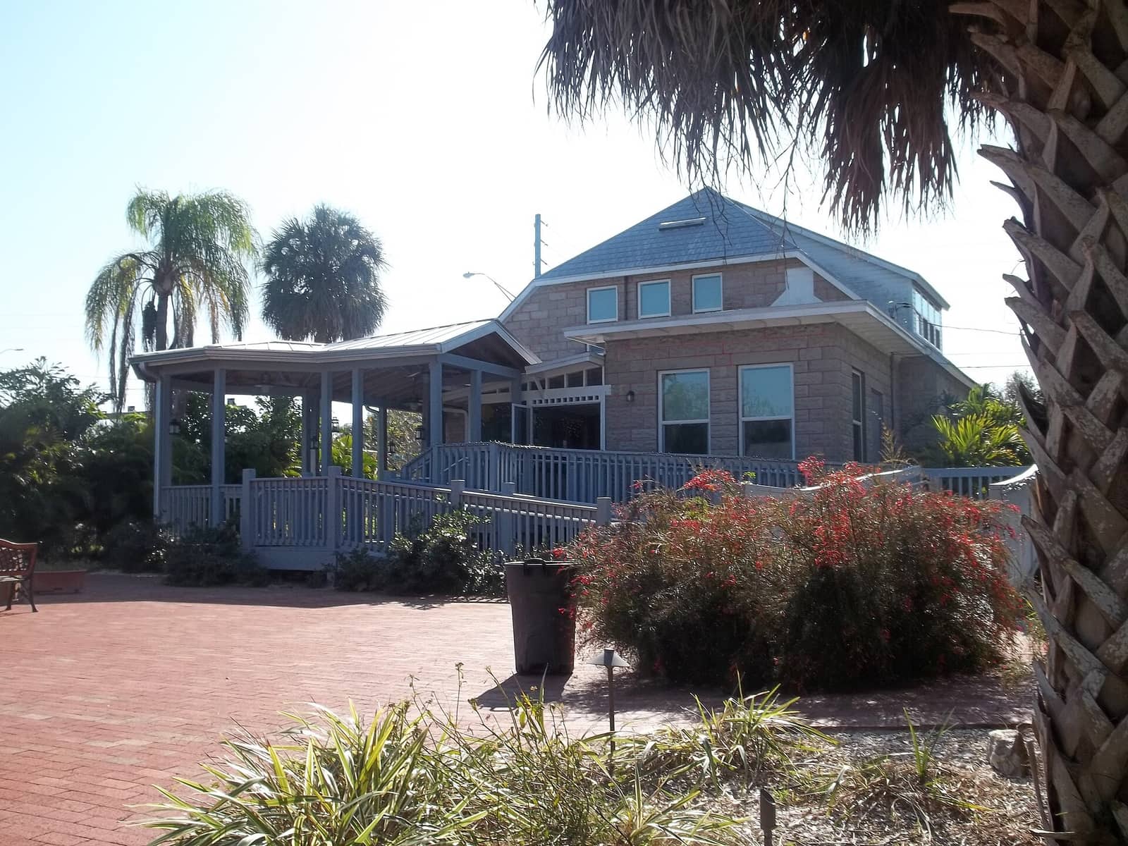 Two-story brick museum building with a wraparound porch, palm trees, and a red brick walkway in front.