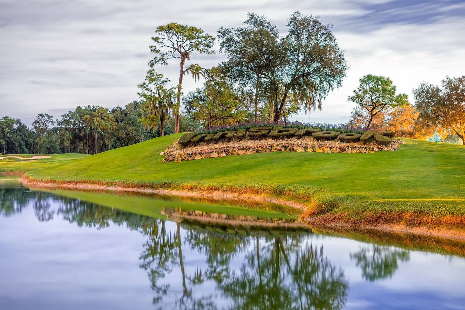 Golf course green with a stone-edged hillside, a pond with reflections, and tall trees under a cloudy sky.