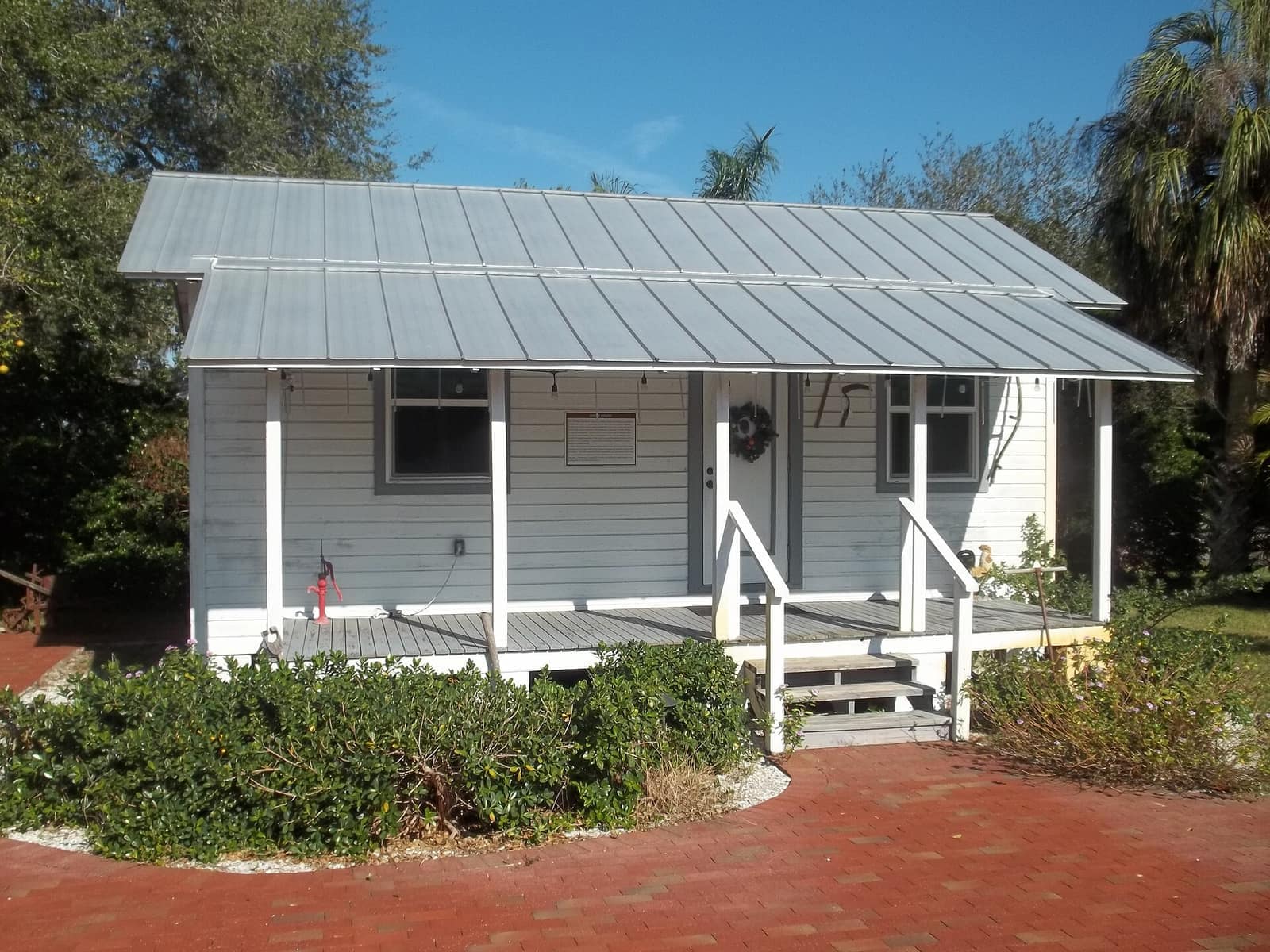 Small white wood cottage with a metal roof, front porch, and wreath on the door, surrounded by shrubs.