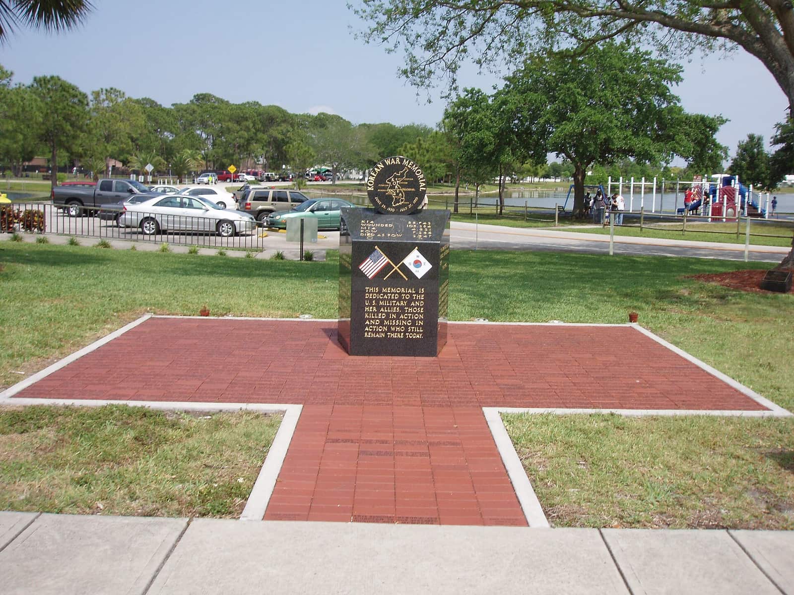 Brick-paved memorial plaza with a dark stone marker, cars and trees behind.