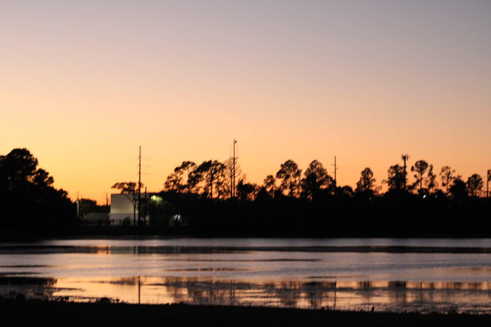 Orange sunset sky reflected on a calm lake with silhouetted trees and a small building on the shore.