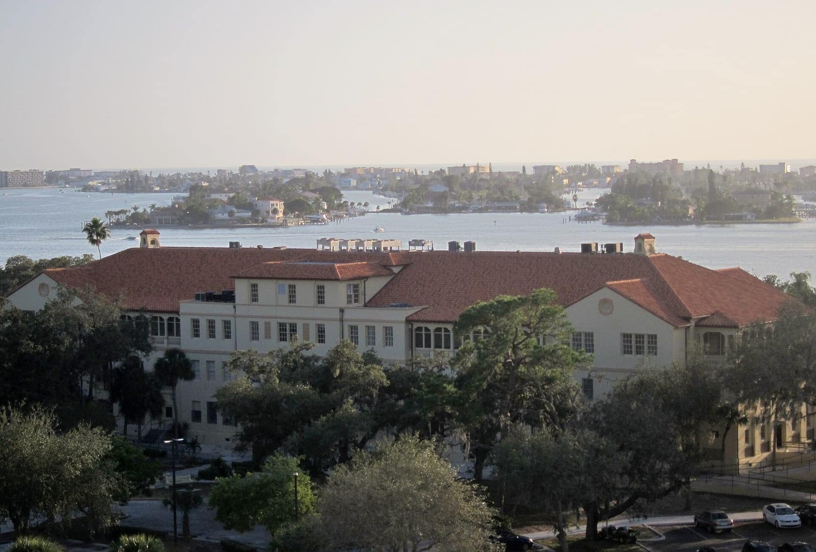 Cream-colored building with red tile roofs overlooks a bay with small islands and houses at dusk.