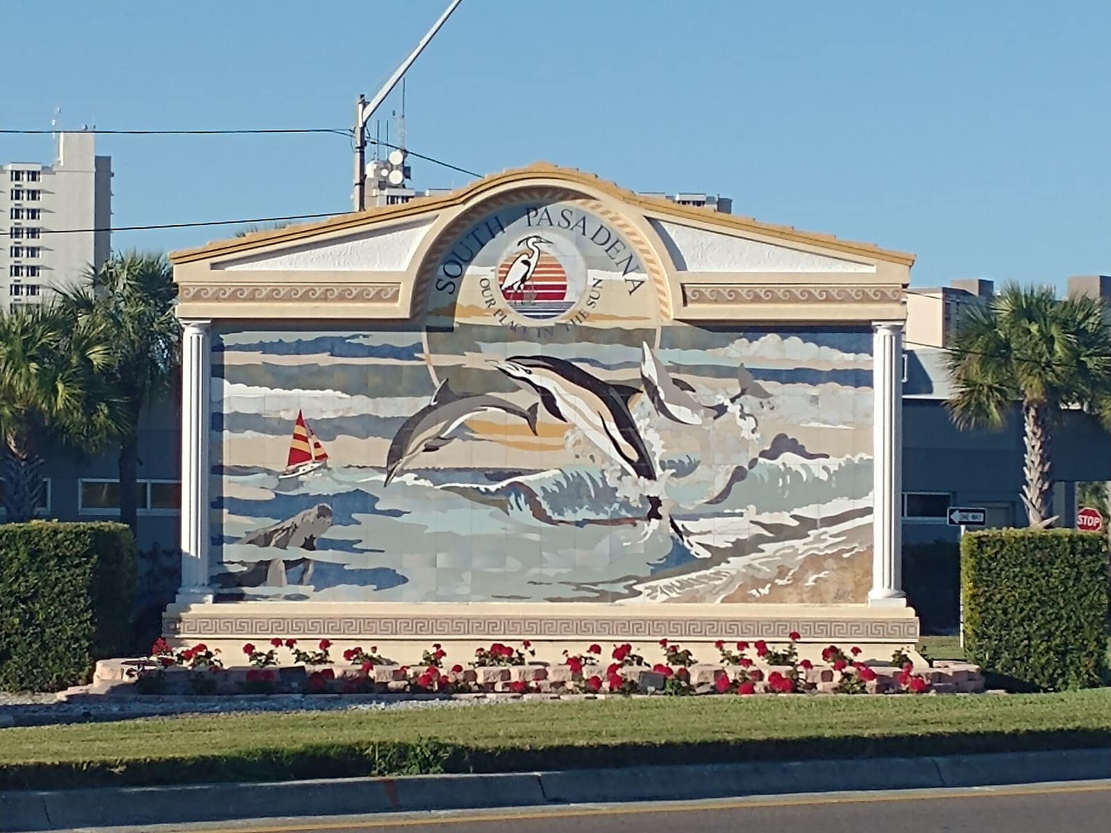 Roadside monument sign with a dolphin and sailboat mural and red flowers along its base.