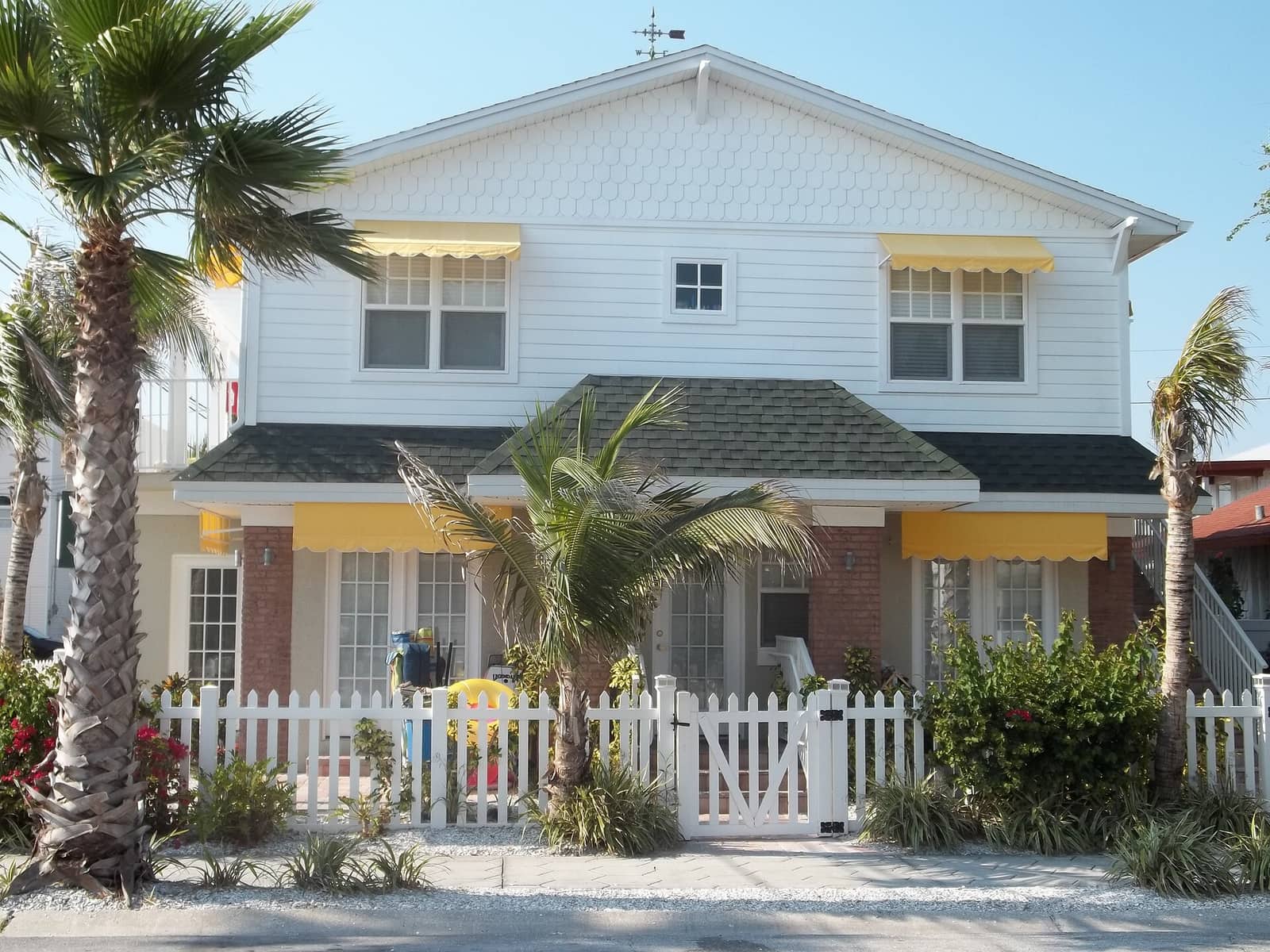 Two-story white house with yellow awnings, brick base, white picket fence, and palm trees in front.