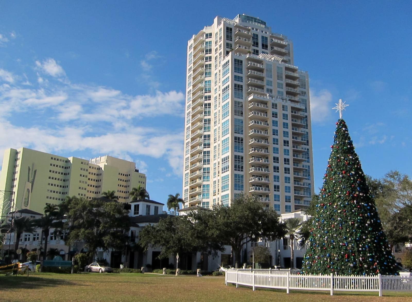 A tall glass condominium tower rises behind a large decorated outdoor Christmas tree on a sunny day.