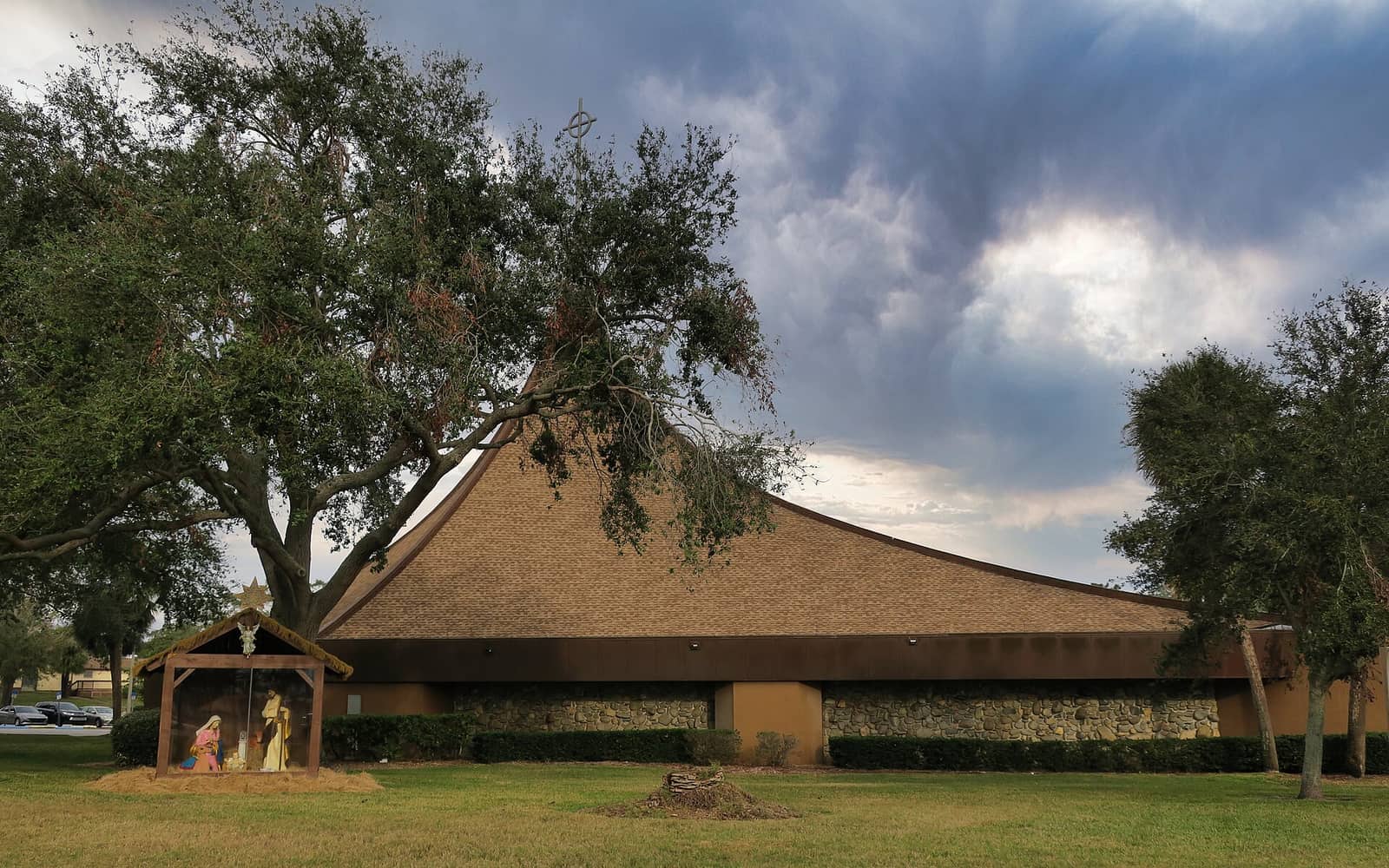A long church building with a wide sloped roof beside a small outdoor nativity scene under cloudy sky.