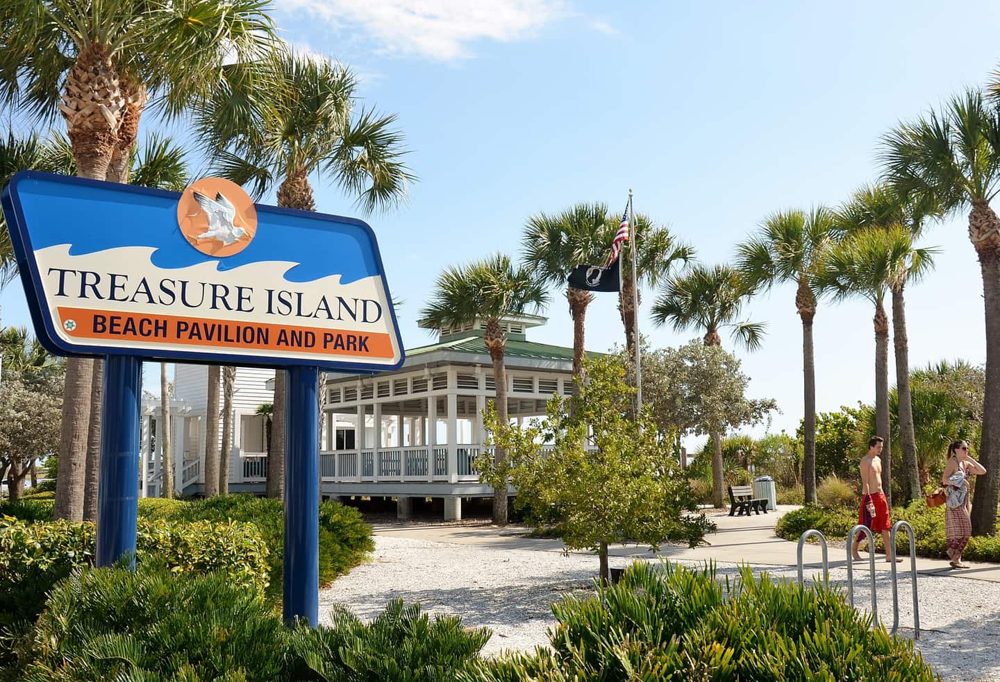 A Treasure Island beach park sign stands beside palm trees, a white pavilion, and a walking path.