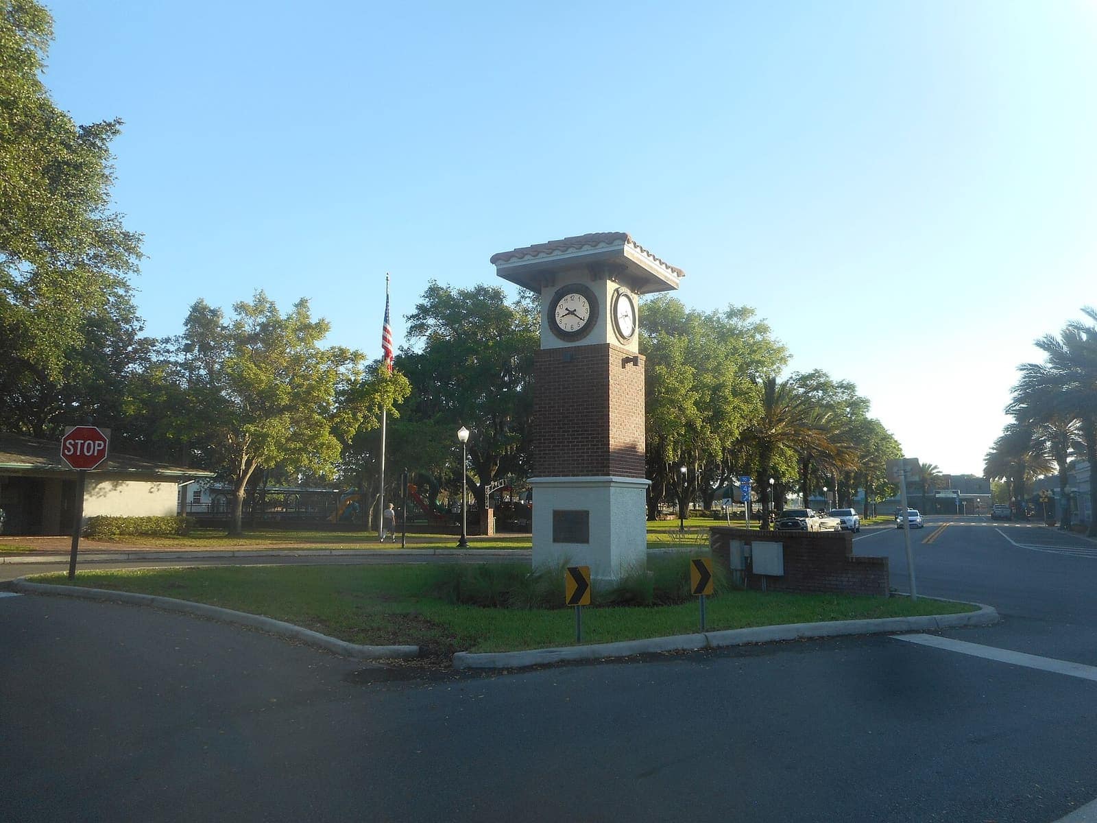 Brick and white clock tower with flagpole and stop sign on a green plaza under a clear morning sky.