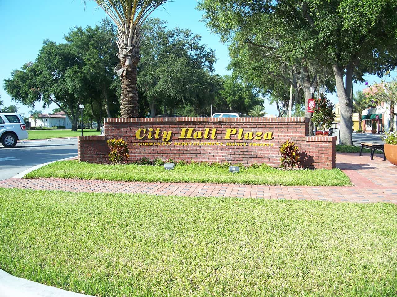 Brick wall sign reading City Hall Plaza in gold letters, with a palm tree, green lawn and brick walkway.