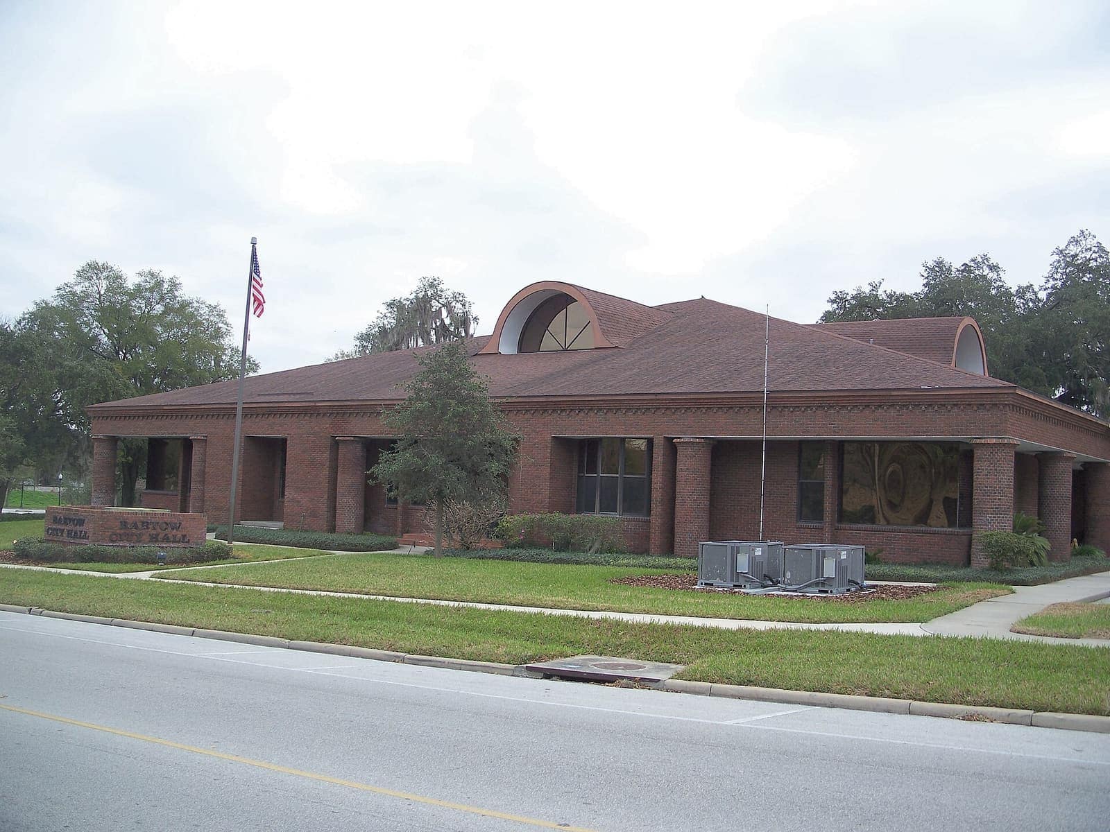 Single-storey brick city hall with a flagpole, arched roof vents and a lawn beside a road.