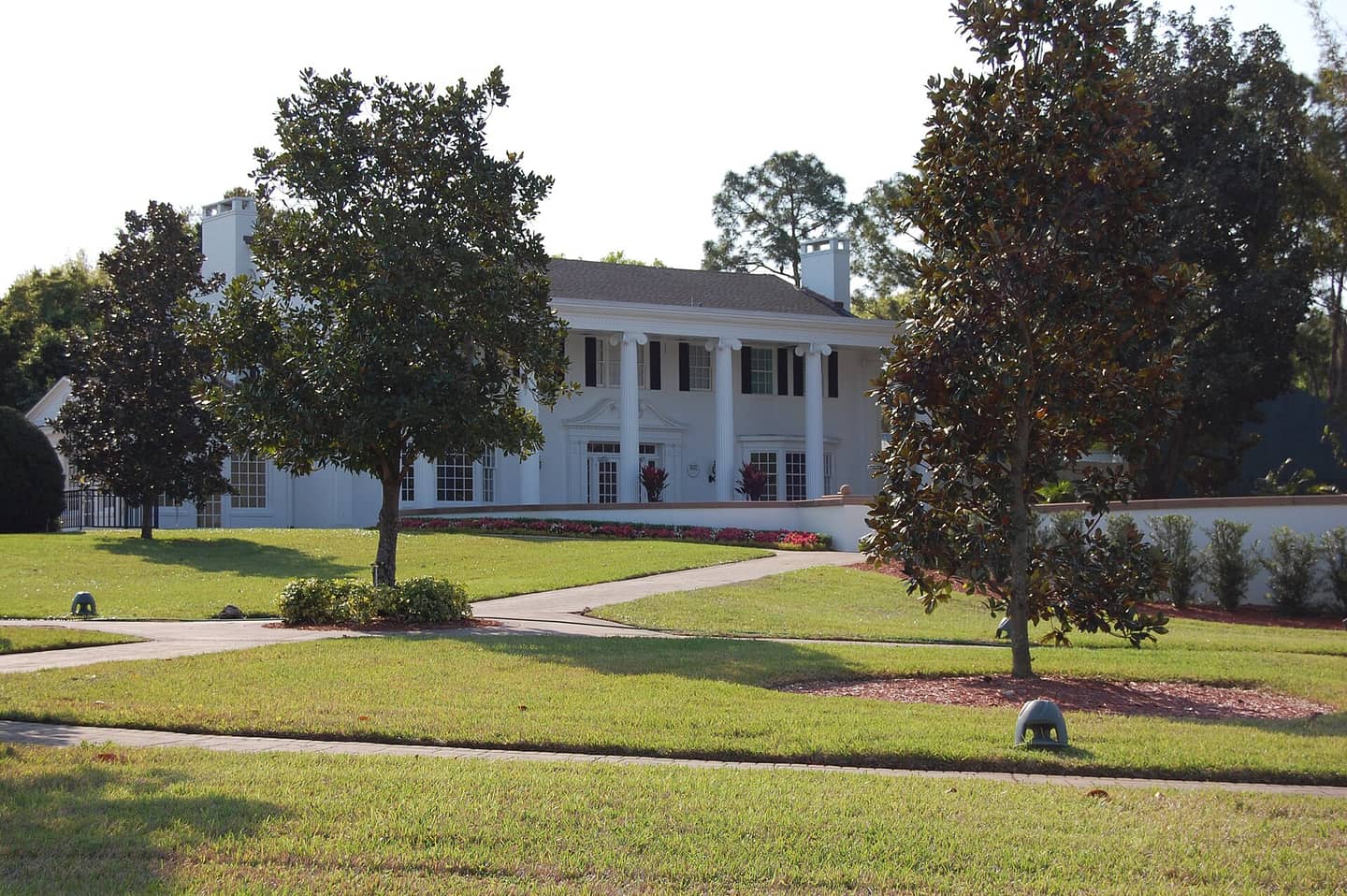 A large white columned house with chimneys stands behind green lawns, a magnolia tree, and a curved walkway.