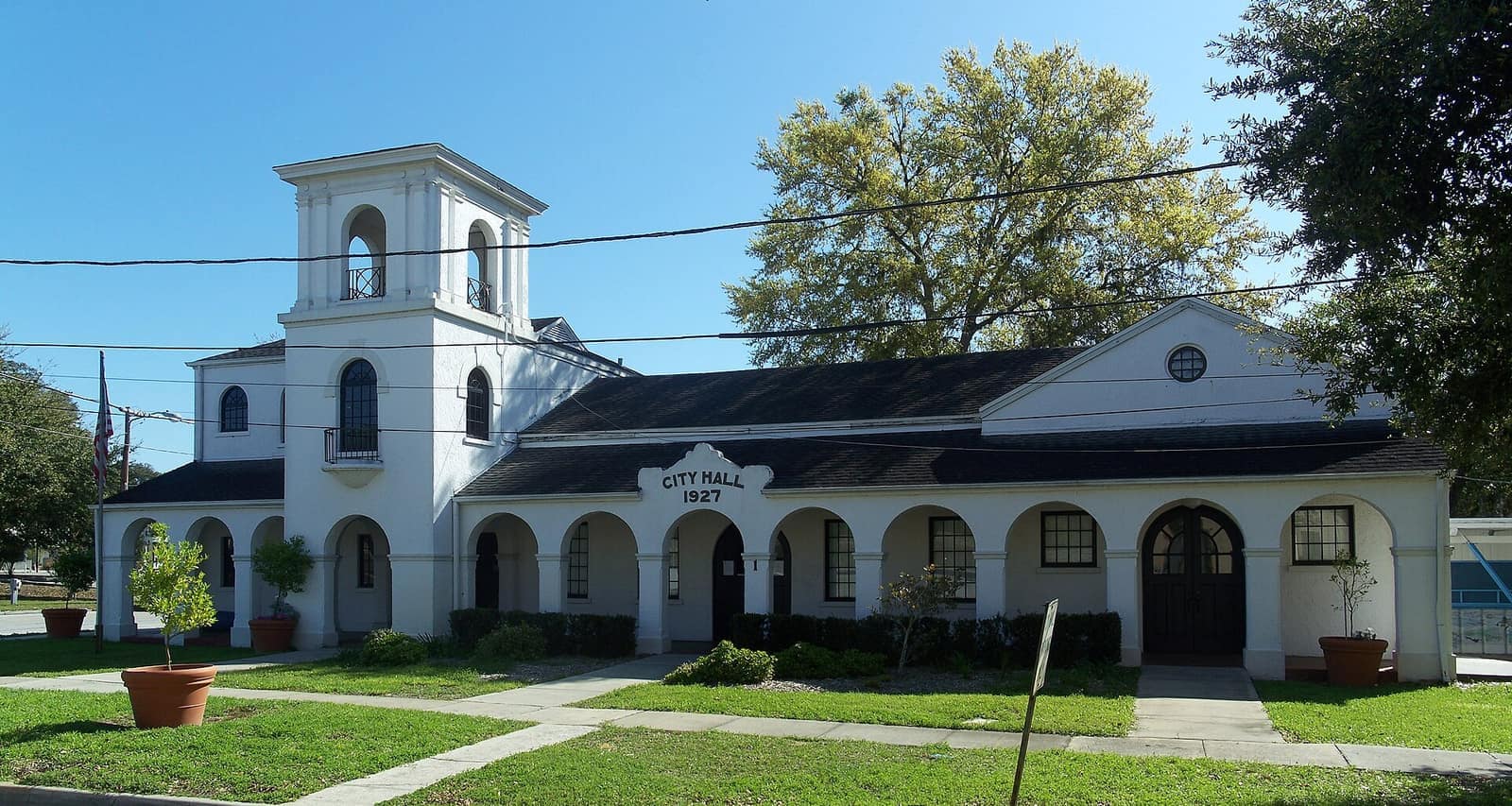 White stucco city hall with bell tower and arched arcade under a clear blue sky.