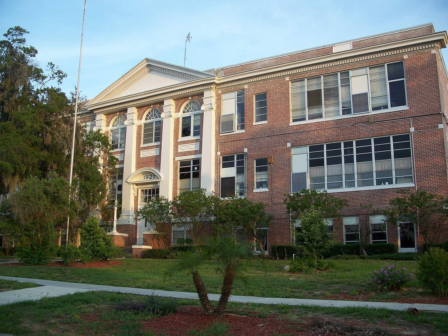 Three-storey brick school building with white columns, large windows and a flagpole beside it.