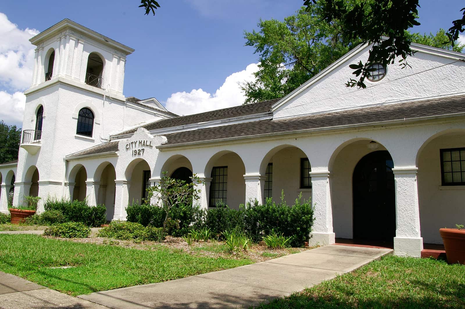 White stucco city hall with arched colonnade, bell tower and potted plants on a sunny day.