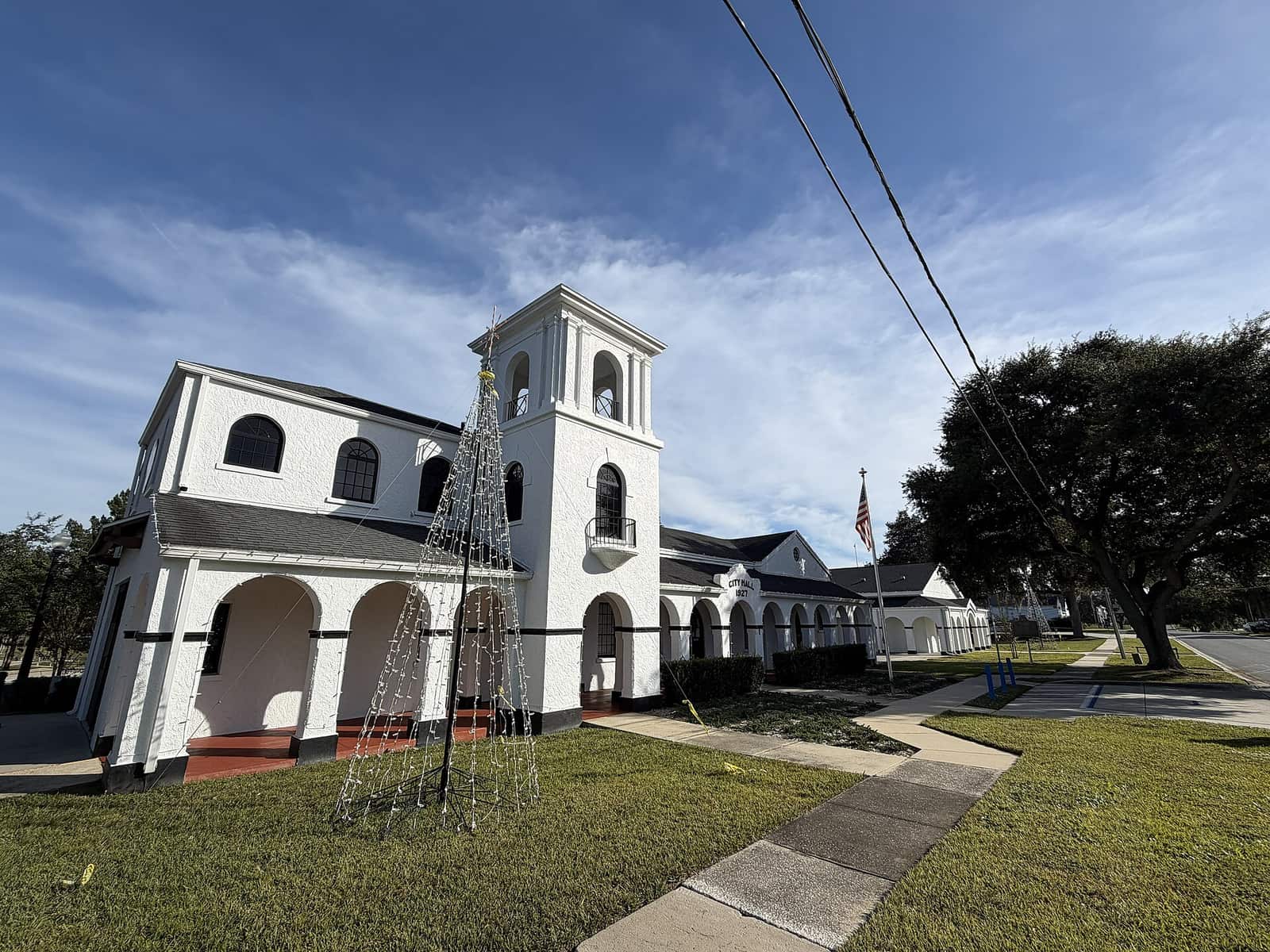 White two-storey city hall with bell tower, arched porch and a lit holiday tree on the lawn.