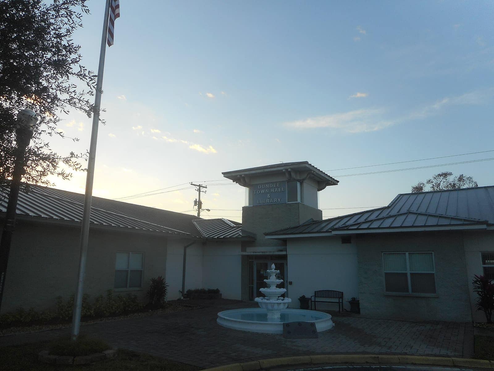 Single-storey light-coloured town hall with a tower, flagpole, and fountain in front under a dusk sky.