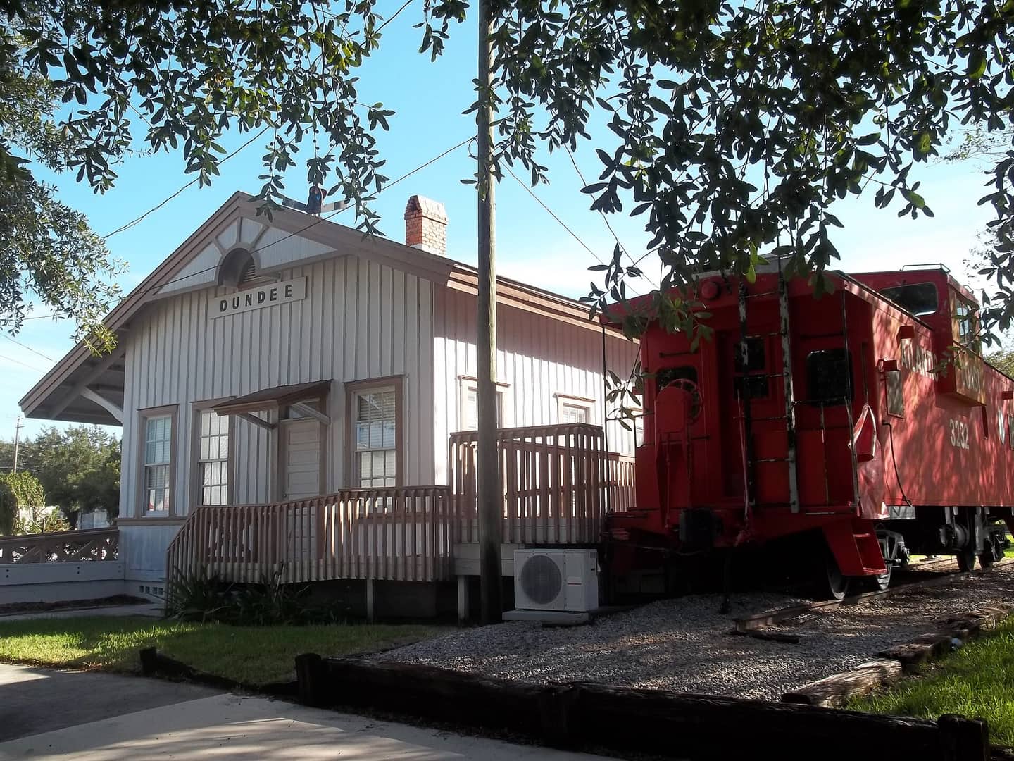 White wooden railroad depot with a red caboose on gravel under trees on a sunny day.