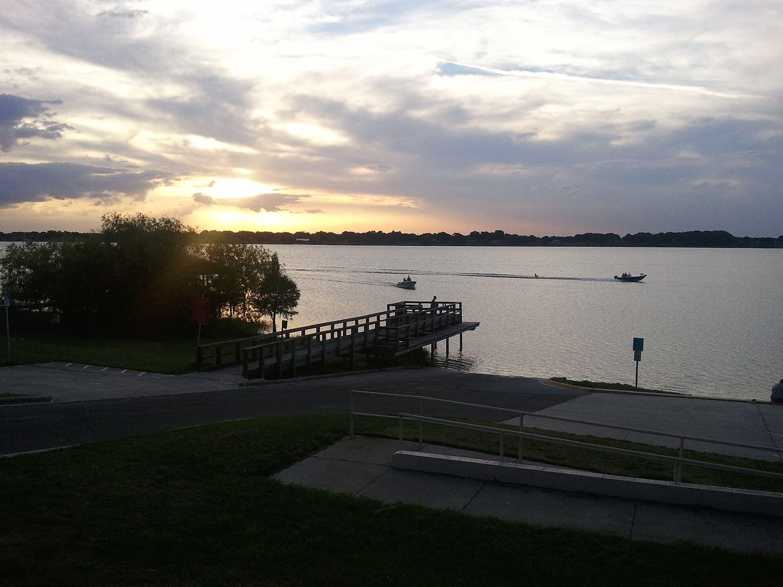 Wooden public boat dock on a calm lake at sunset, with two small boats and trees along the shore.