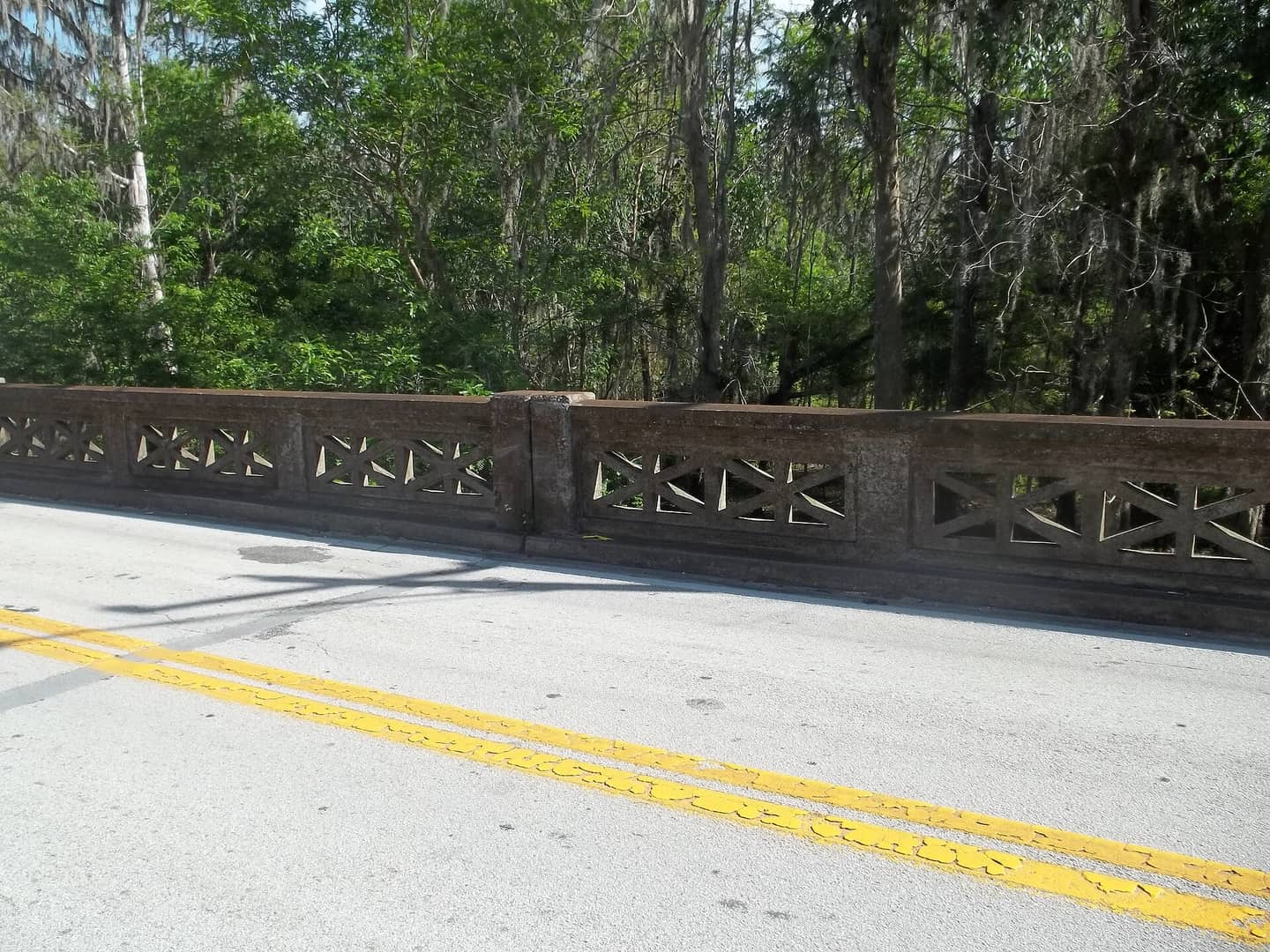 Close view of an ornate pierced concrete bridge railing beside a paved road with trees behind.