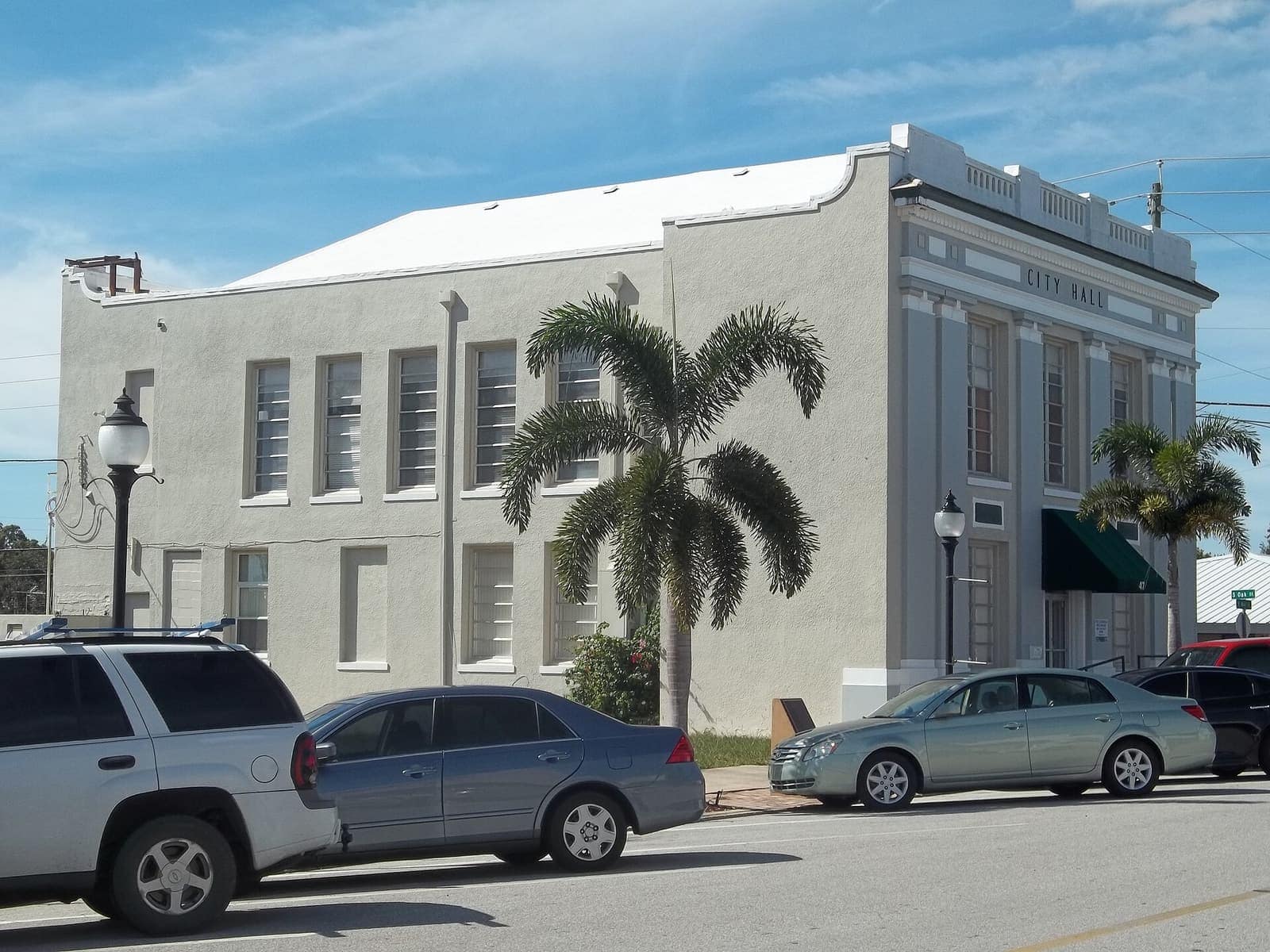 White two-story former city hall with columns and a green awning beside parked cars under blue sky.