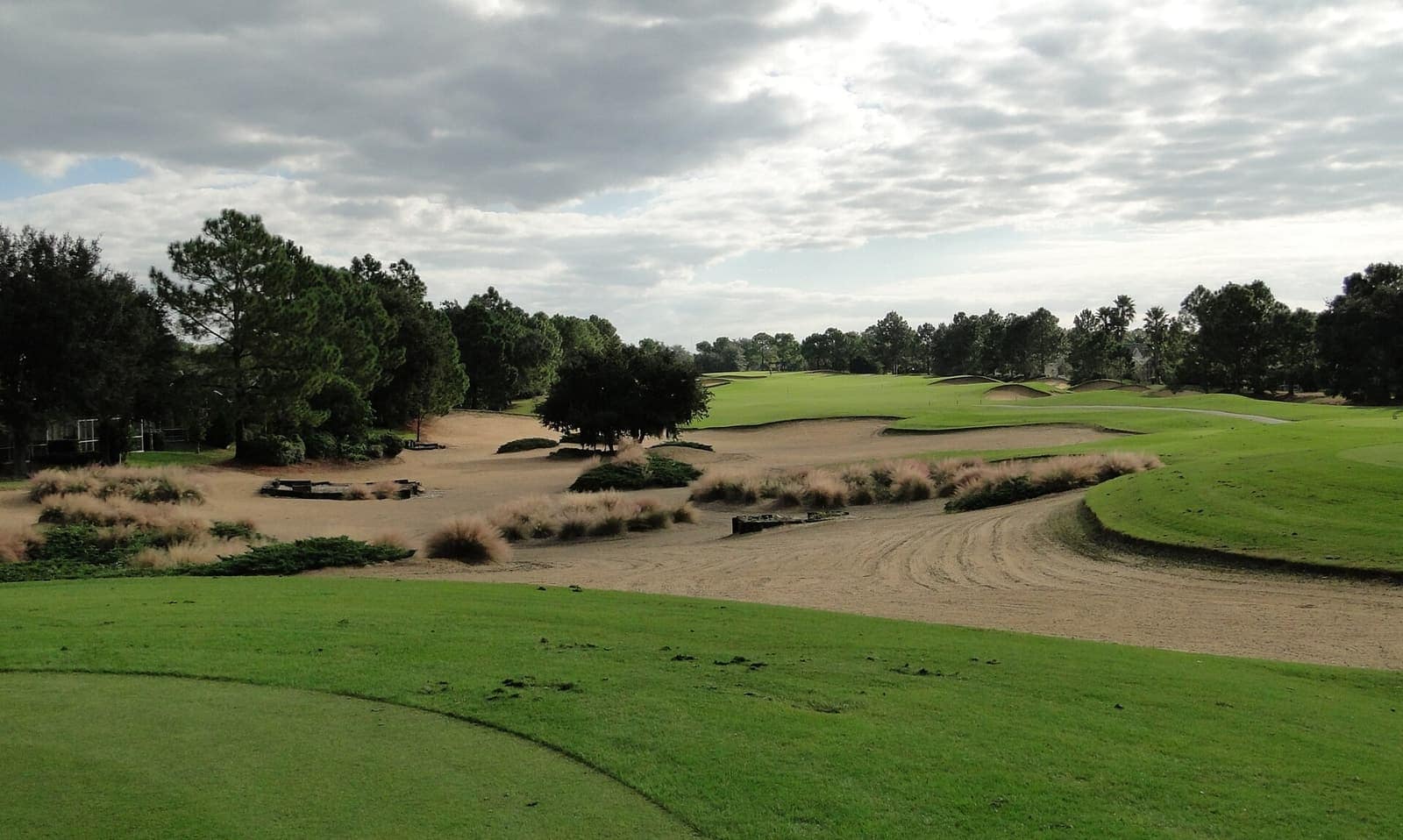 Golf course fairway with sand bunkers, grass mounds, and pine trees under a partly cloudy sky.