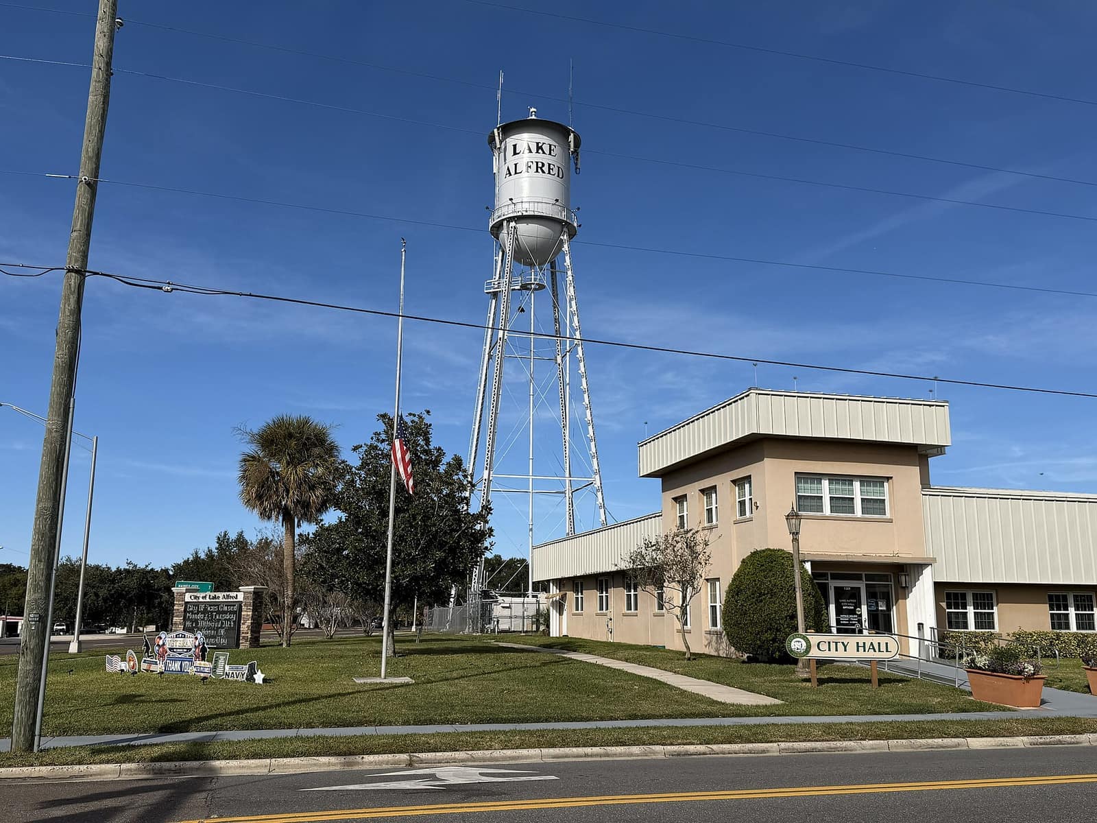 Tan city hall building beside a white water tower with a flag, under a clear blue sky on a sunny day.