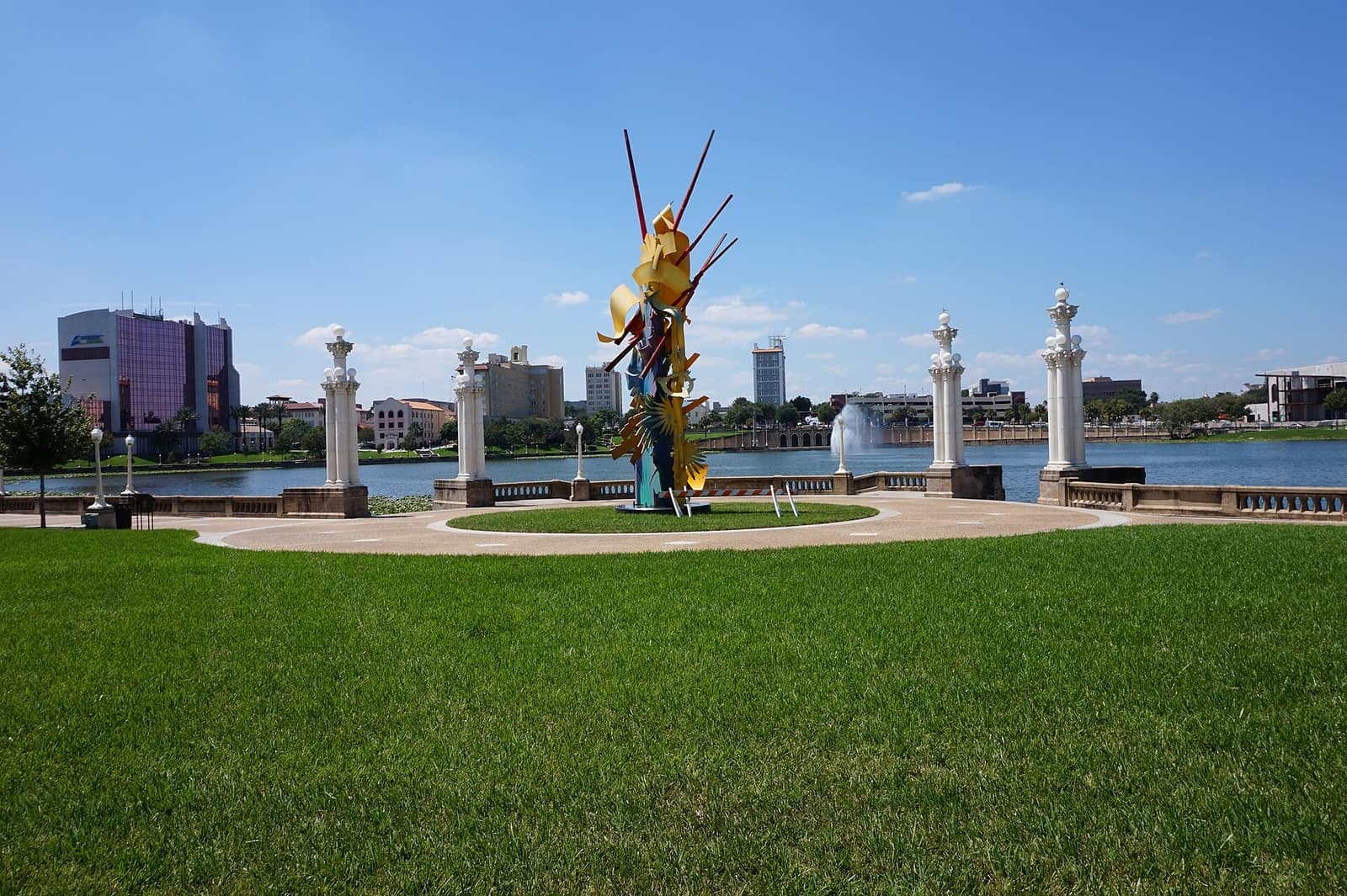 Colorful abstract metal sculpture stands in a grassy park beside a lake, fountain, and distant buildings.