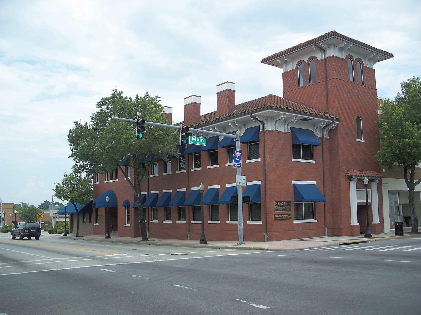 Two-story red brick corner building with a tower, blue awnings, and a traffic signal on a city street.